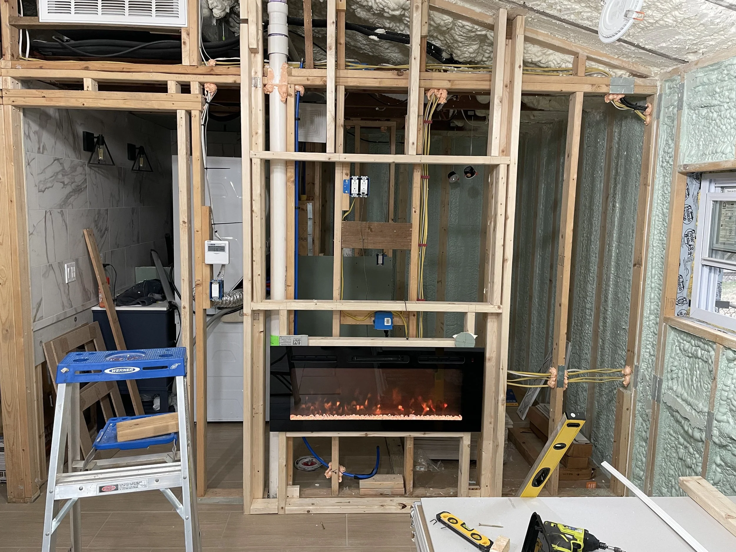 Interior of a house under construction with exposed wooden framing, electrical wiring, insulation, and a fireplace, tools and equipment visible around the room.
