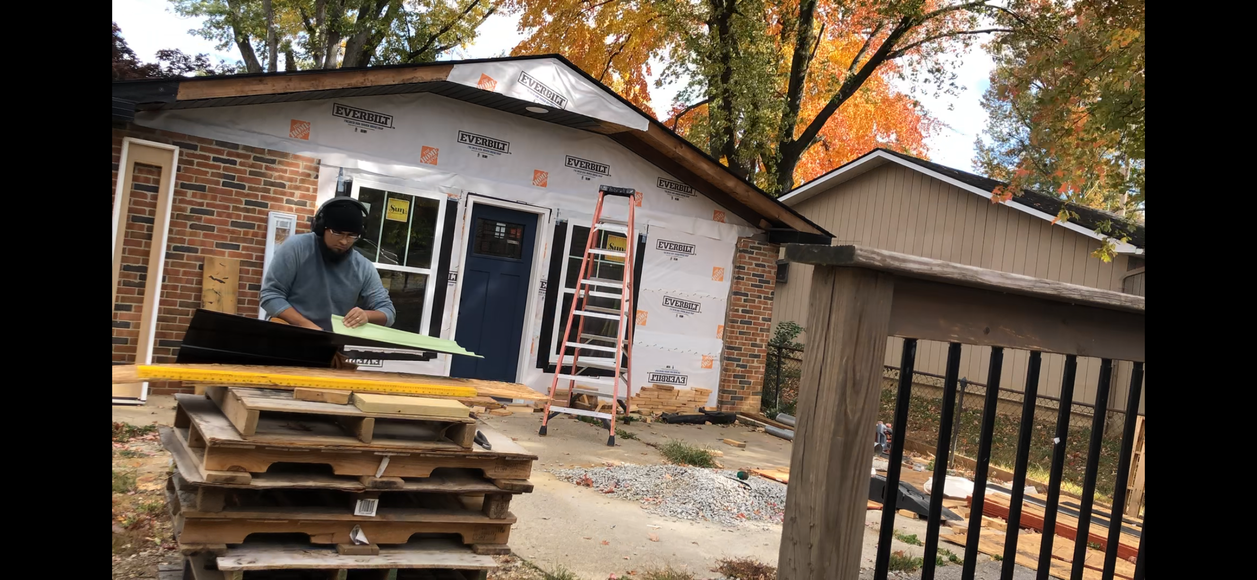 A person wearing headphones working on a house renovation, with construction materials and scaffolding outside a house with brick and siding, under autumn trees.