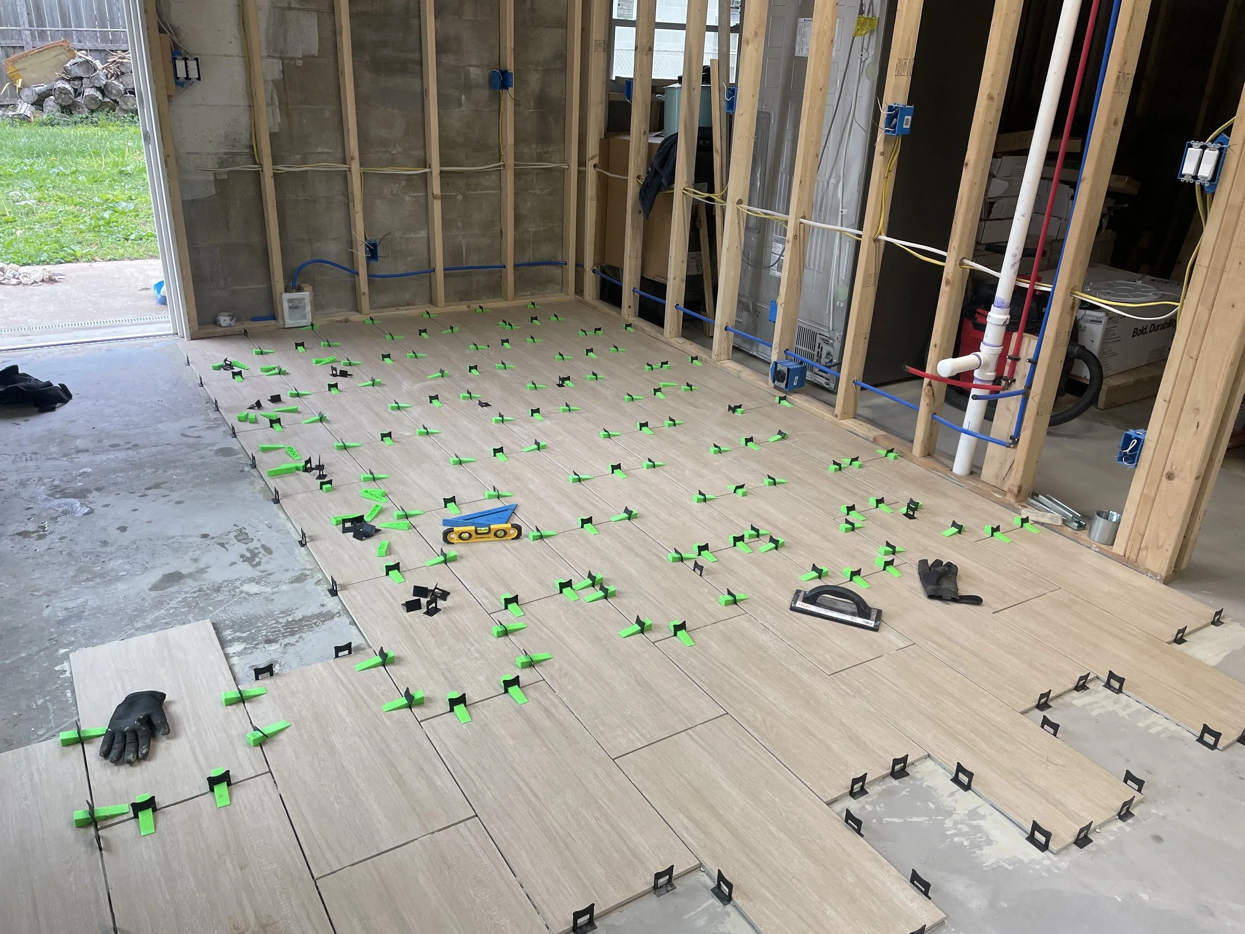 Interior of a house under construction with wooden framing, electrical wiring, and a floor being tiled with green tile spacers and tools visible.