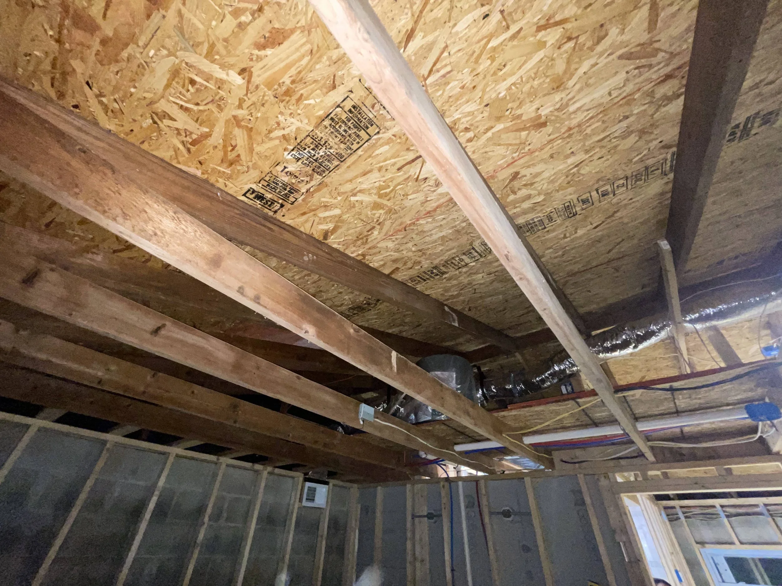Interior view of a building under construction with exposed wooden framing, OSB sheathing on the ceiling, and visible HVAC ductwork and electrical wiring.