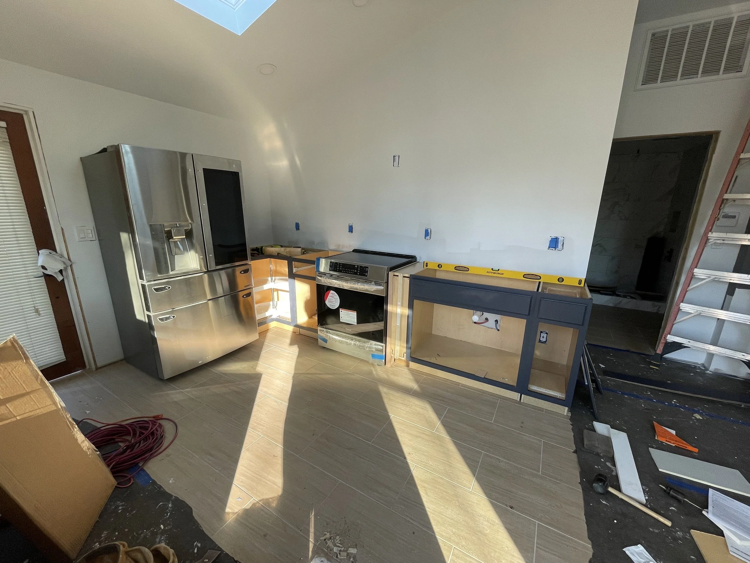 A partially constructed kitchen showing a stainless steel refrigerator, a stove, unfinished cabinets, and an open wall with blue electrical outlets. There are tools, a ladder, and construction materials scattered around.