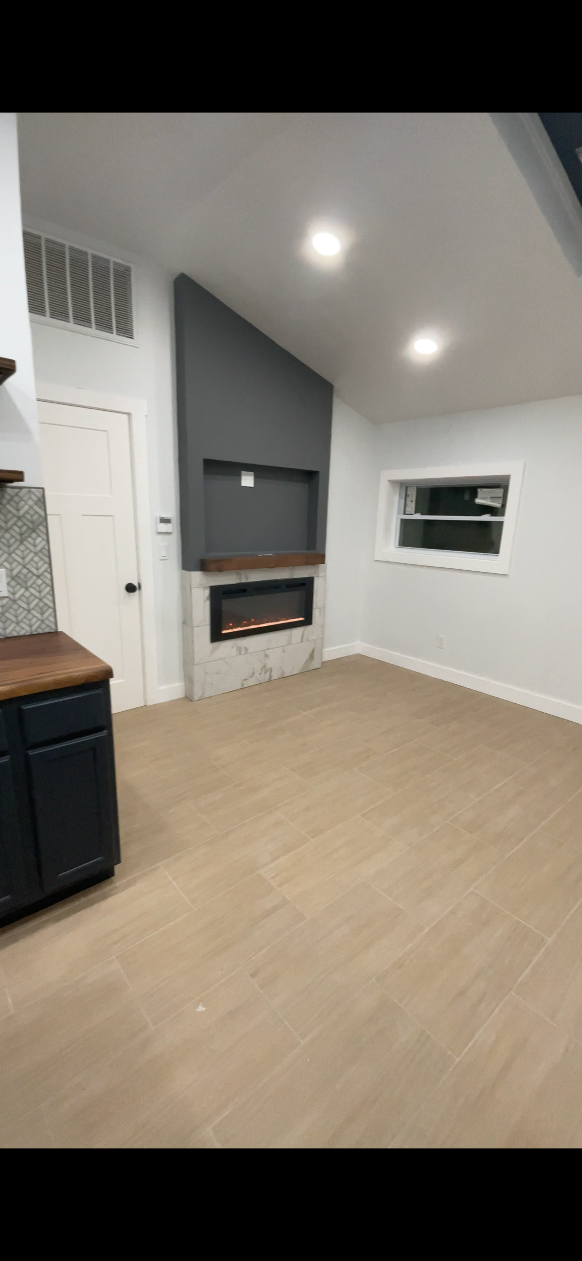 Living room with beige tiled flooring, gray accent wall with a fireplace, and a small window, illuminated by ceiling lights.
