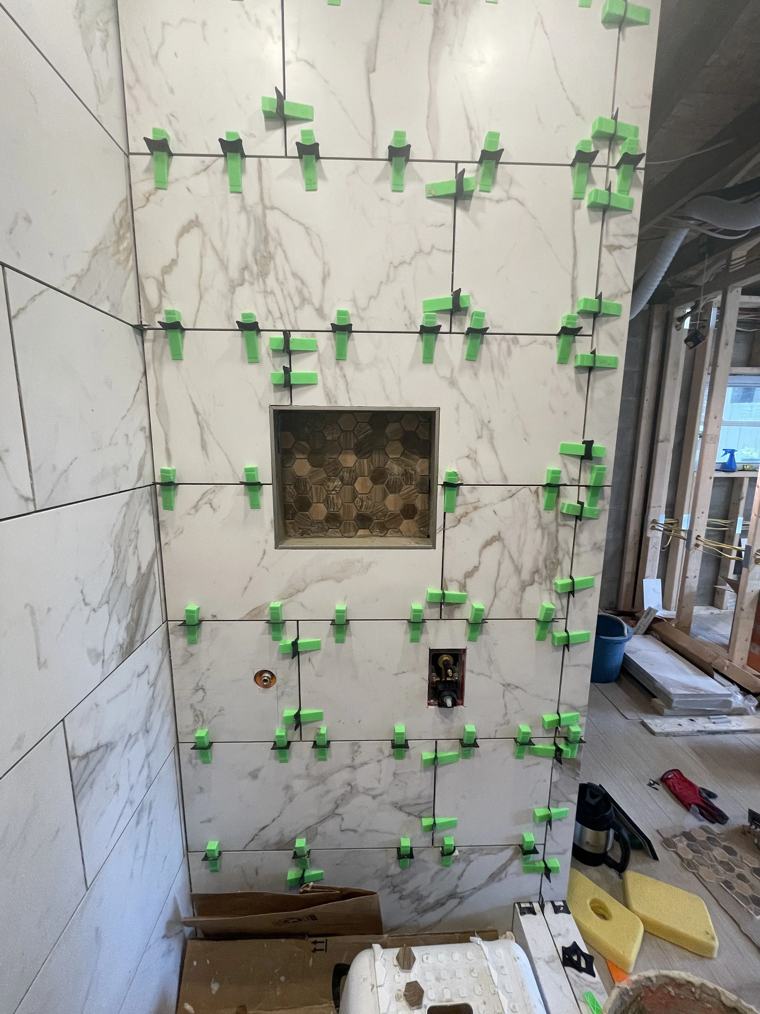 Unfinished bathroom wall with marble tiles and black spacers, small open square niche with decorative tiles, construction tools and supplies on the floor.