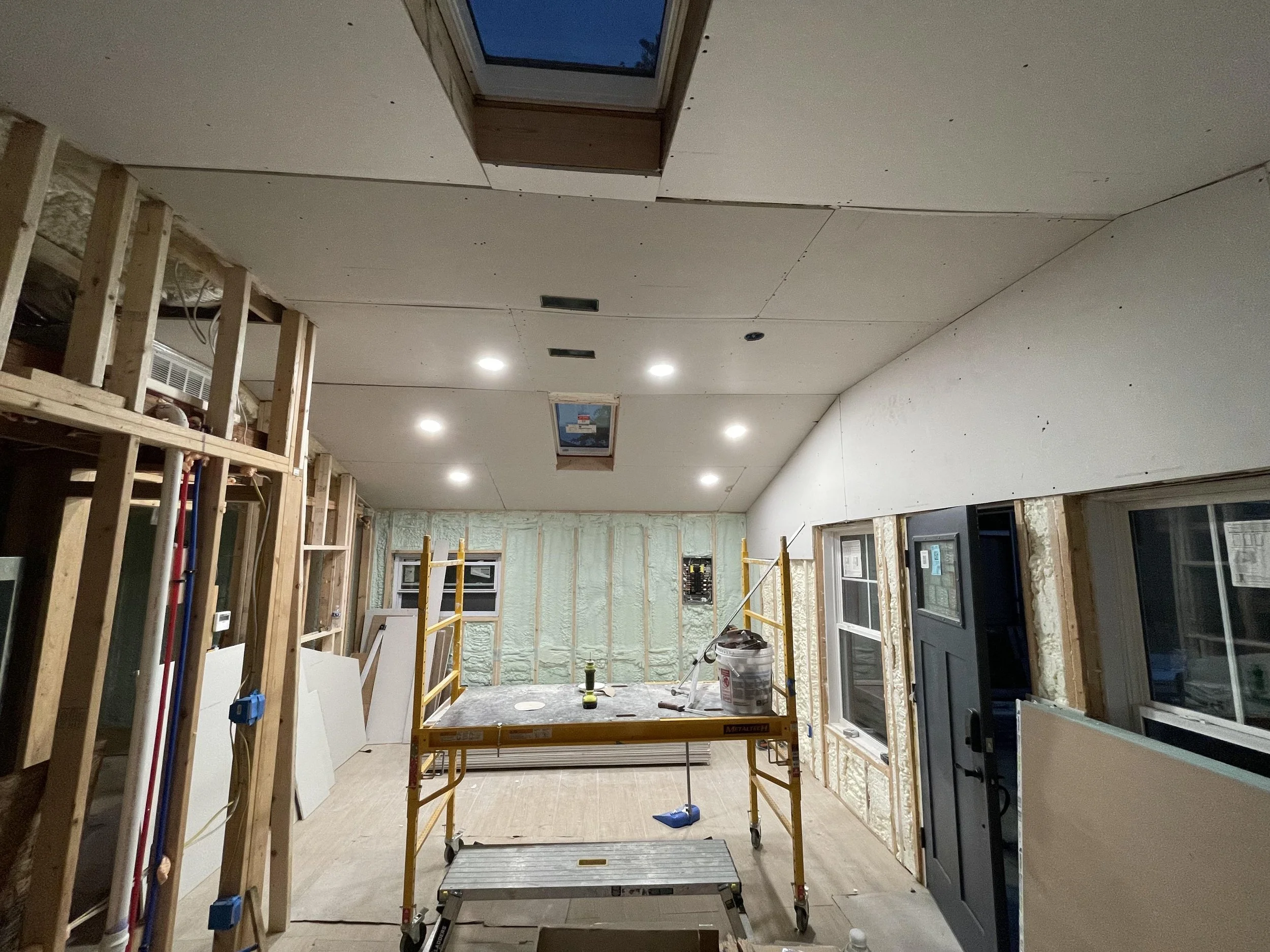 Interior of a room under construction, with exposed wooden framing, drywall installation in progress, and various construction tools and materials scattered around.