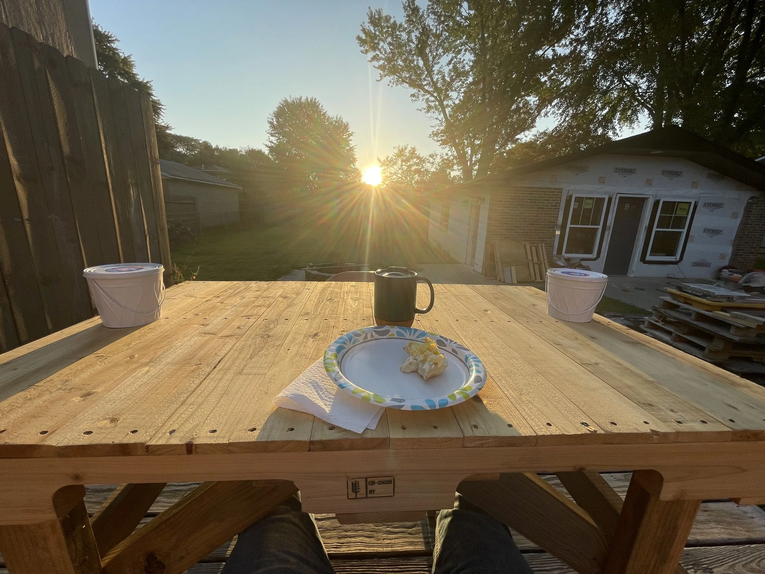 A wooden deck with a plate of scrambled eggs, two cups, and a black mug, overlooking a backyard with a house under construction, trees, and the setting sun.