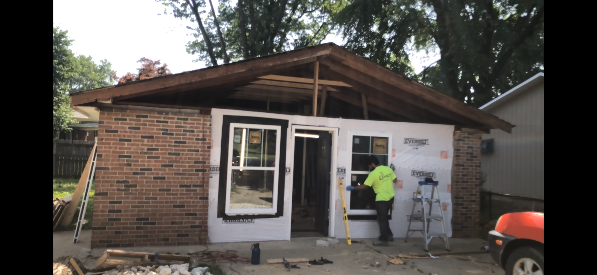 Construction worker installing a window on a house under renovation, with a brick wall and wooden roof structure visible.