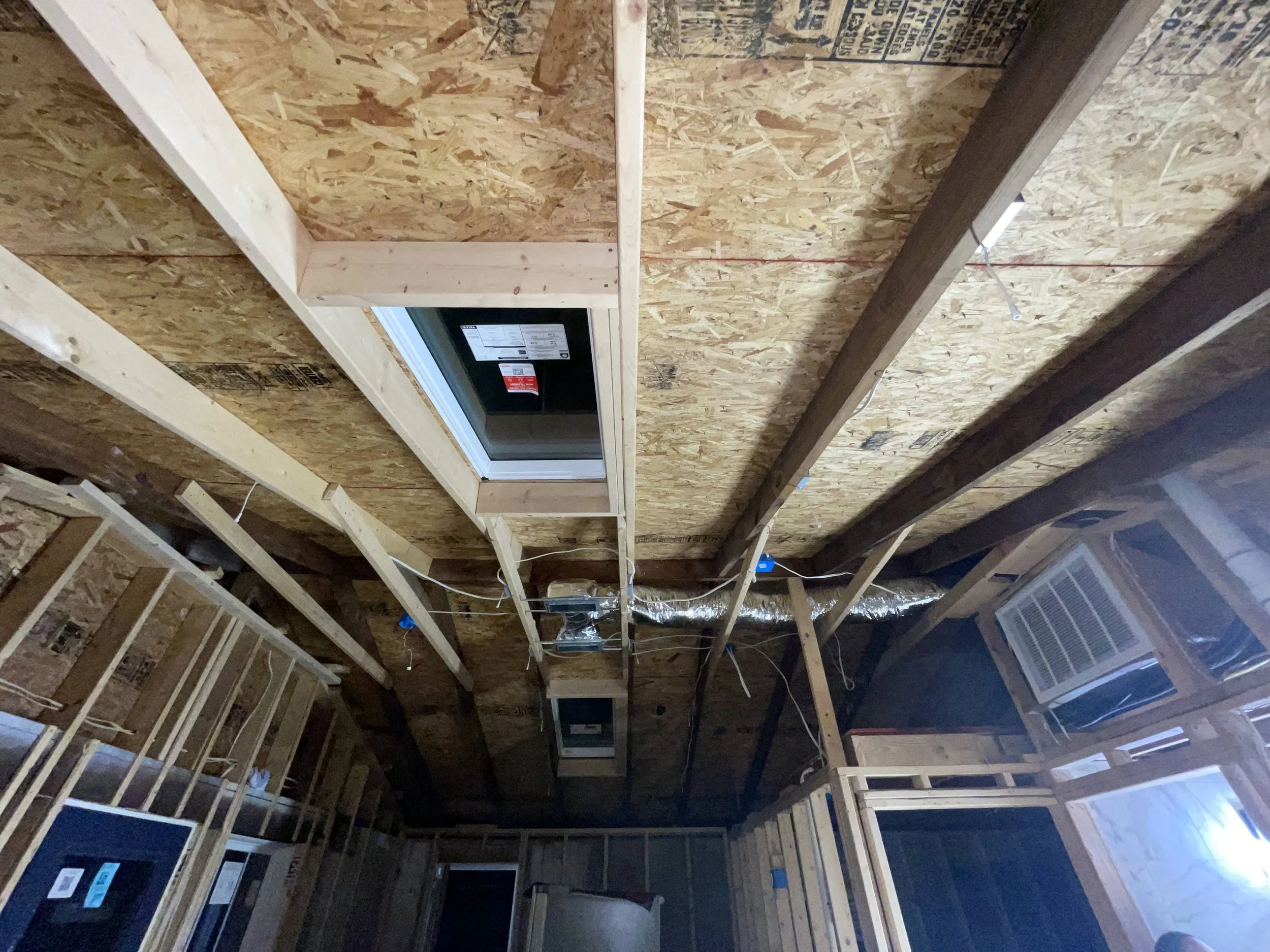 The ceiling of an unfinished room with wooden beams, OSB paneling, and a ceiling vent.