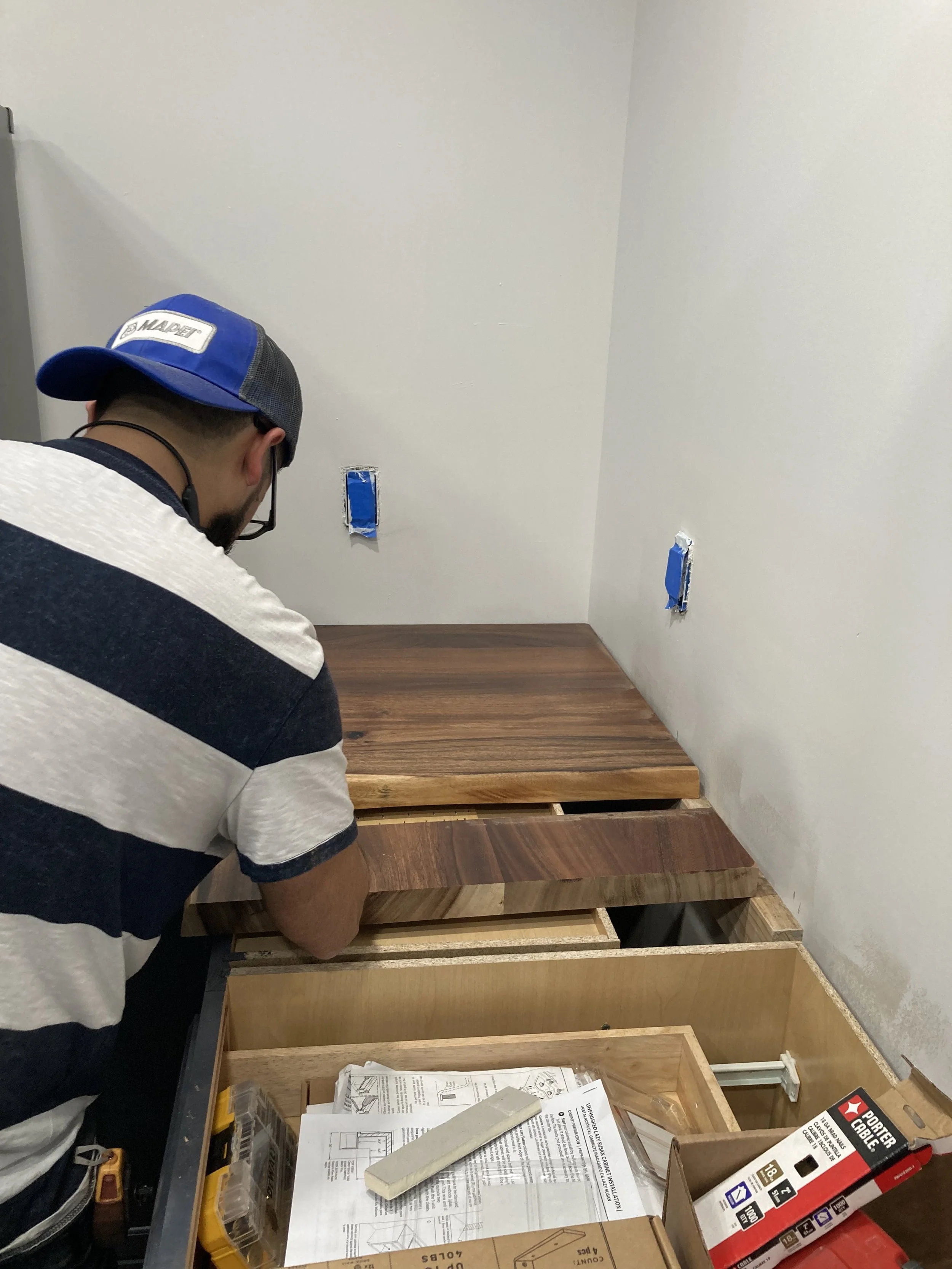 A person wearing a blue and black cap and black glasses is assembling furniture in a room with white walls and wooden floors; there are disassembled furniture pieces, tools, instructions, and packaging materials on the table and on the floor.