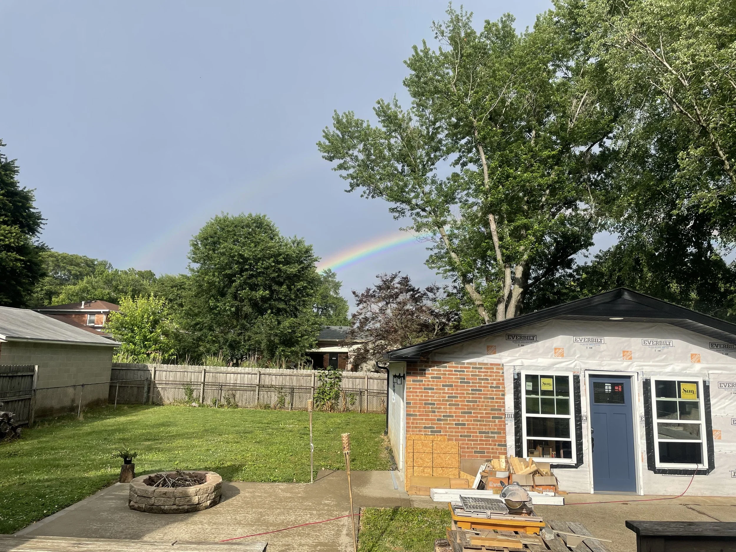 A backyard under construction with a partly built brick house, construction materials, and tools in the yard. Trees and a rainbow are visible in the sky.