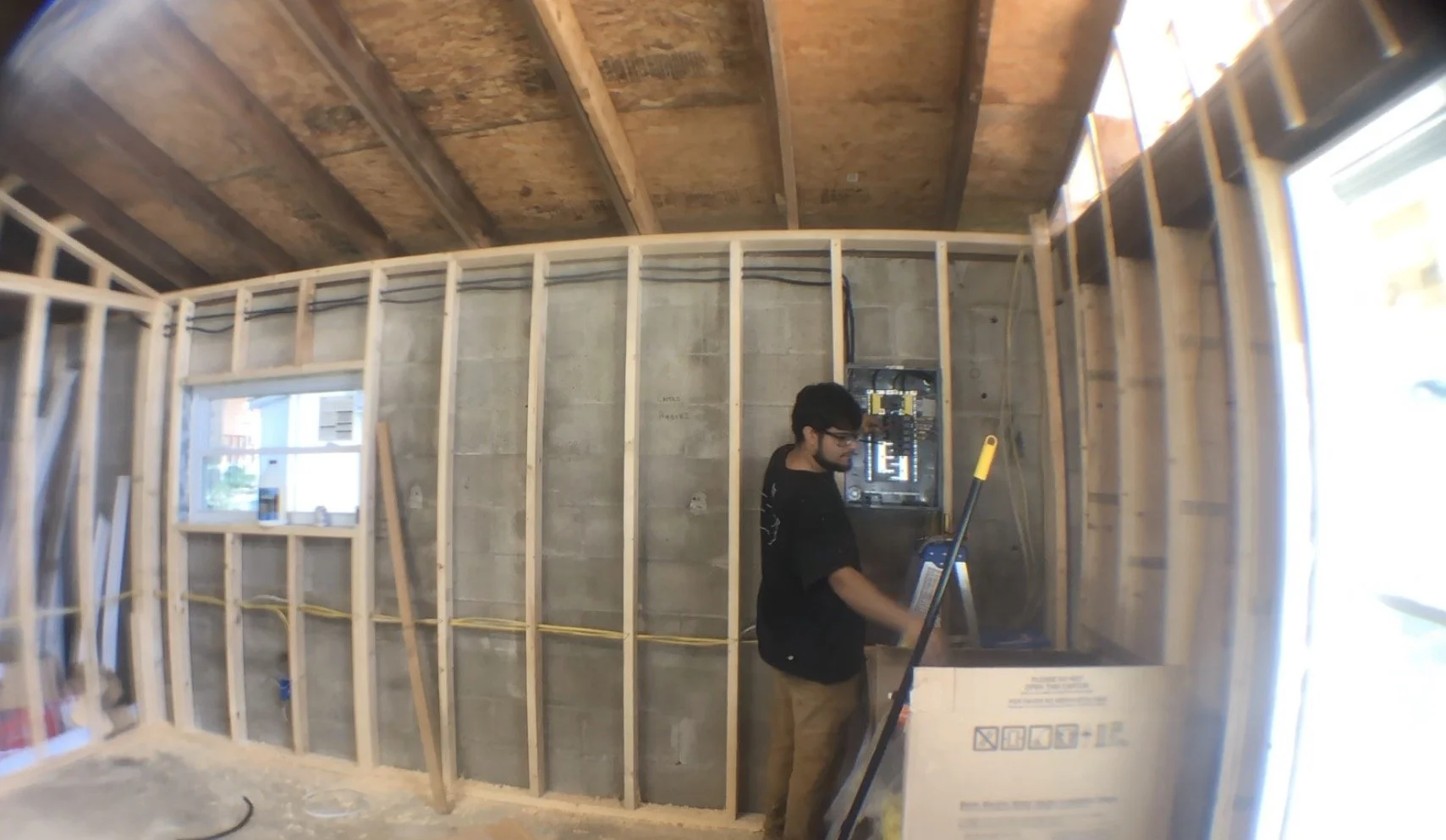 A person working on electrical wiring inside a room under construction, with exposed framing and an electrical panel on the wall.