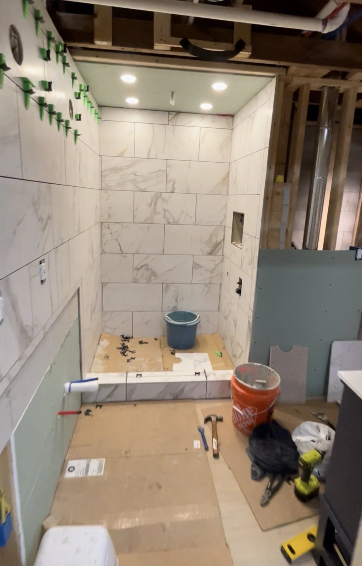 Under renovation bathroom with tiled shower walls and construction tools and materials such as a bucket, screwdrivers, a drill, and drywall on the floor.