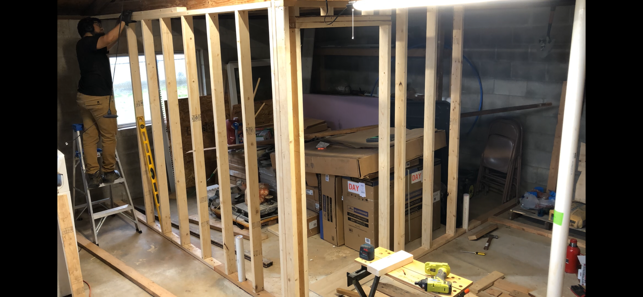 A person on a ladder working on an interior wall framing in a construction project. The room includes stacked boxes, tools, and construction materials.