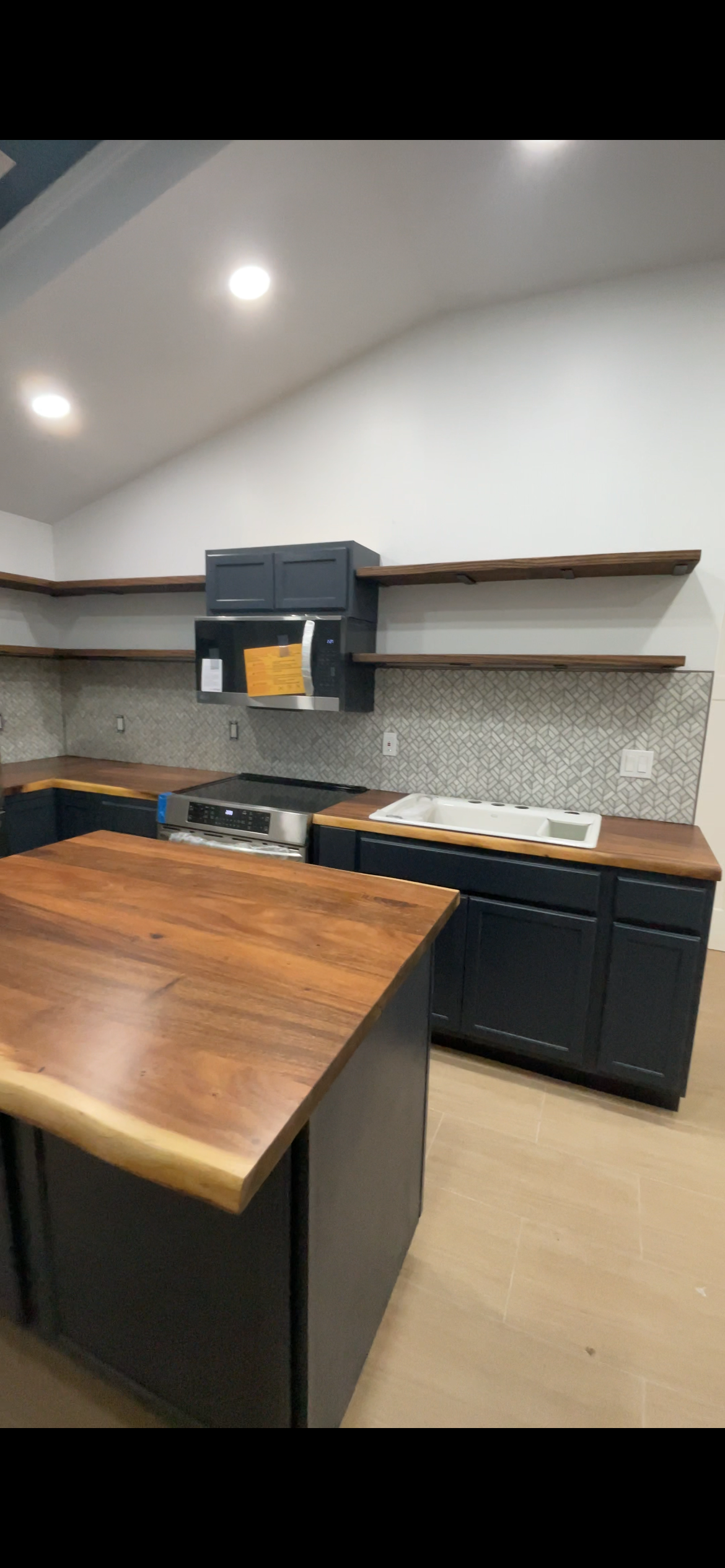 Modern kitchen with wooden countertops, dark blue cabinets, open wooden shelves, and a geometric backsplash. Includes a stove, microwave, and sink.