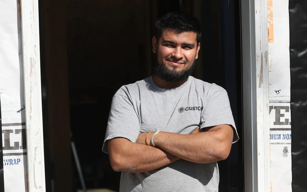 Young man with a beard and short black hair standing at a doorway, smiling with arms crossed, wearing a gray T-shirt
