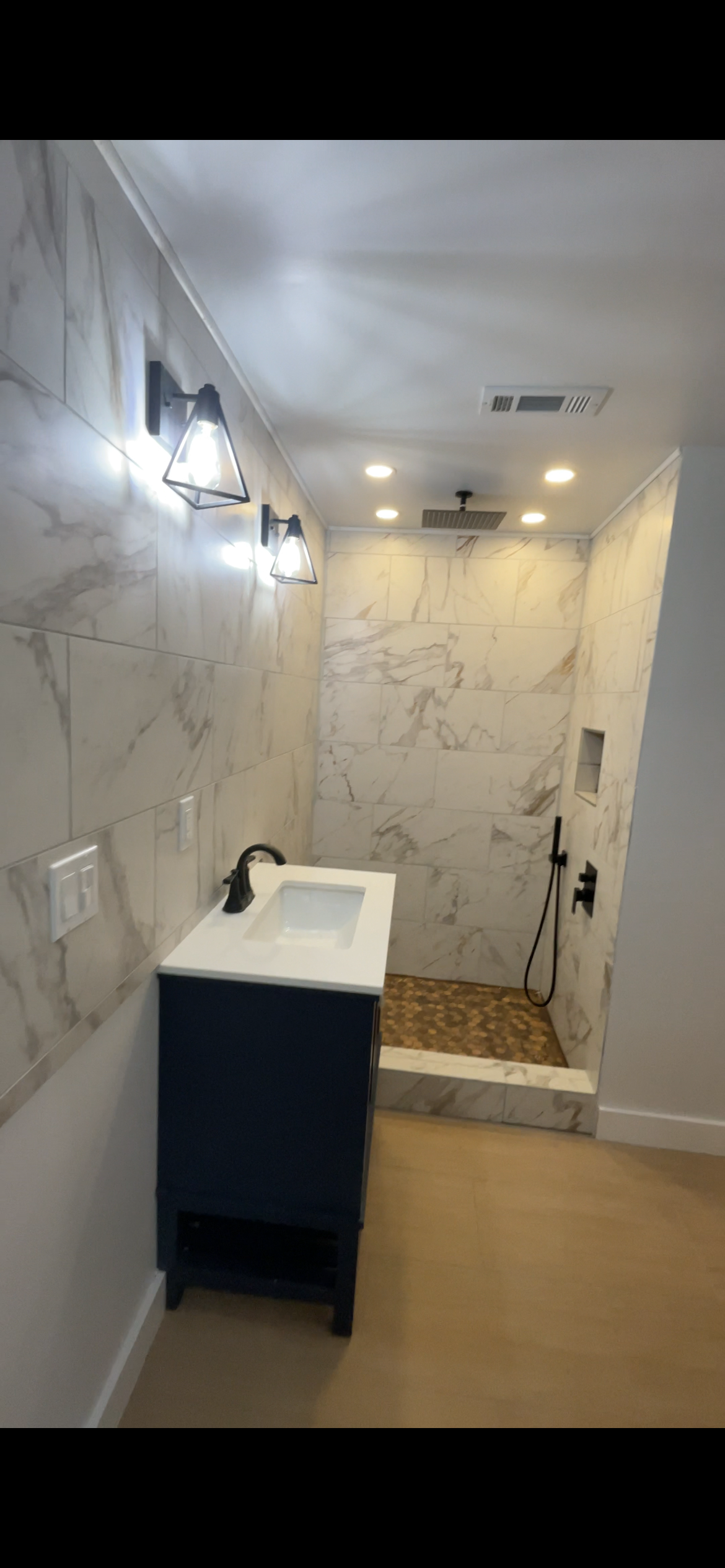 Bathroom with marble walls, a small vanity with a white sink, black fixtures, and a walk-in shower with a pebble floor and black showerhead.