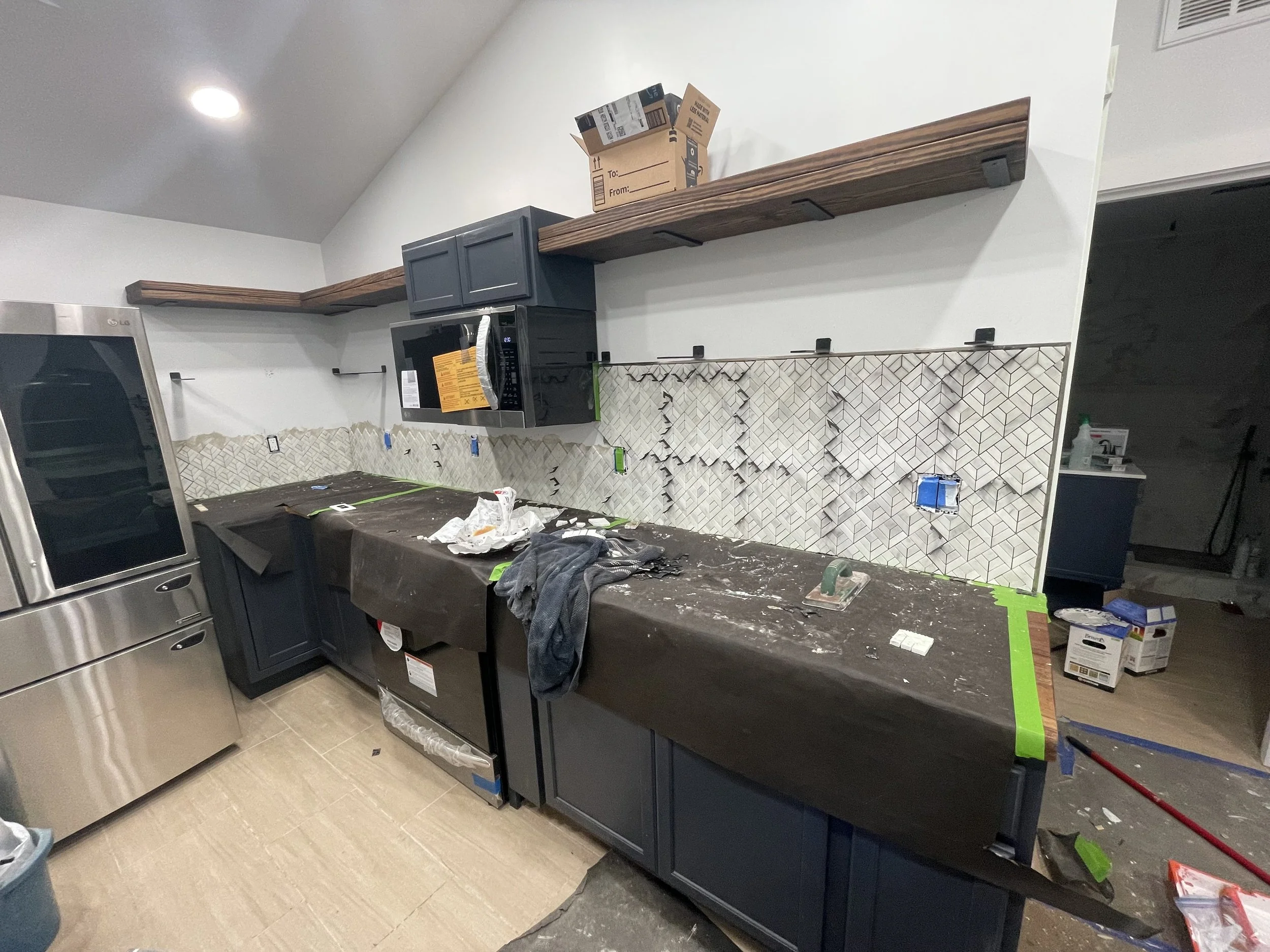 Kitchen in the process of being renovated with dark blue lower cabinets and open wooden shelves. A microwave is installed in the cabinets, and tools and materials are scattered on the black countertop, indicating ongoing work.