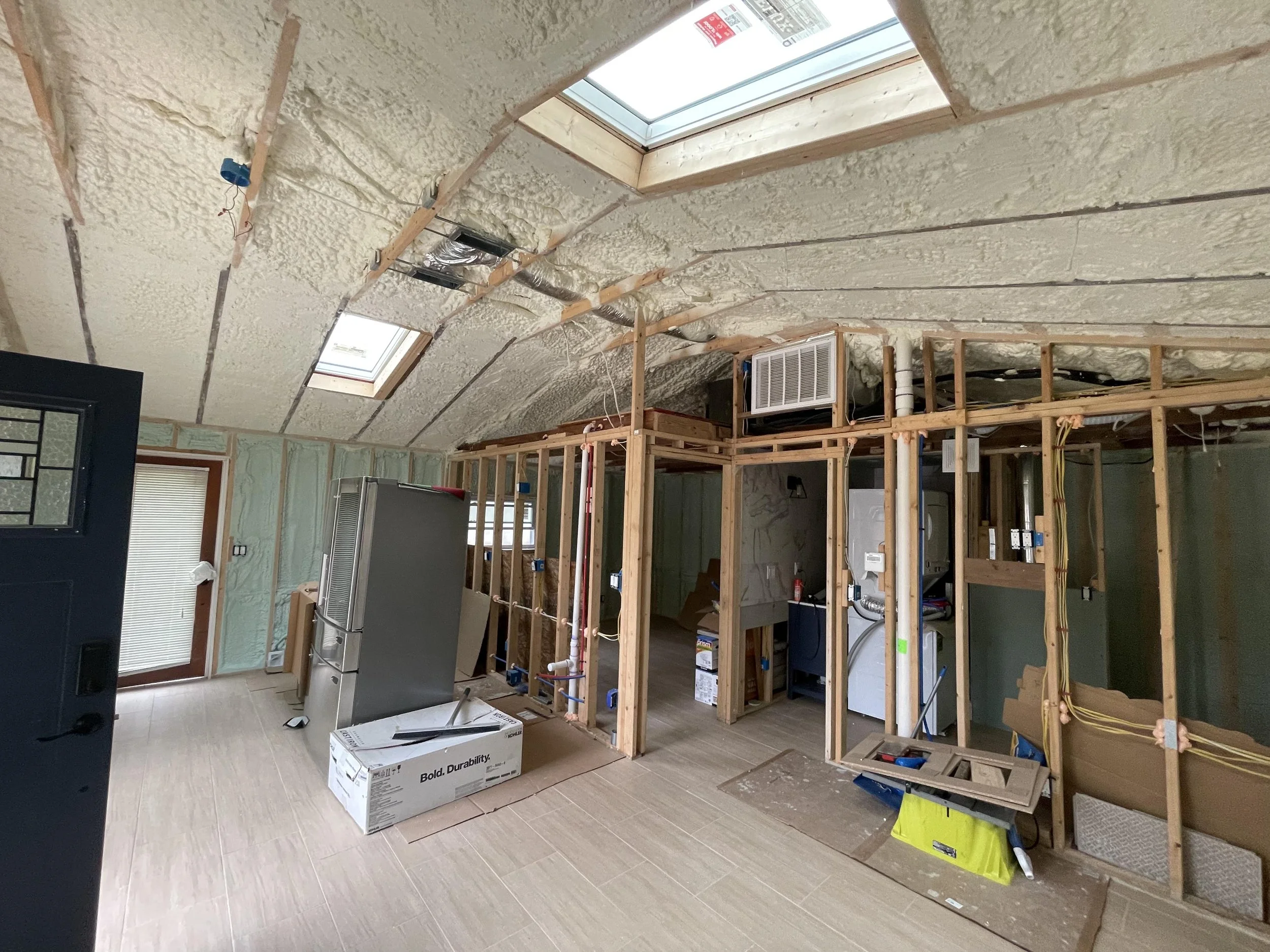 Interior of a house under renovation with partially installed insulation on the ceiling and exposed wooden framing. A skylight window is on the ceiling, and there are various construction materials and tools around.