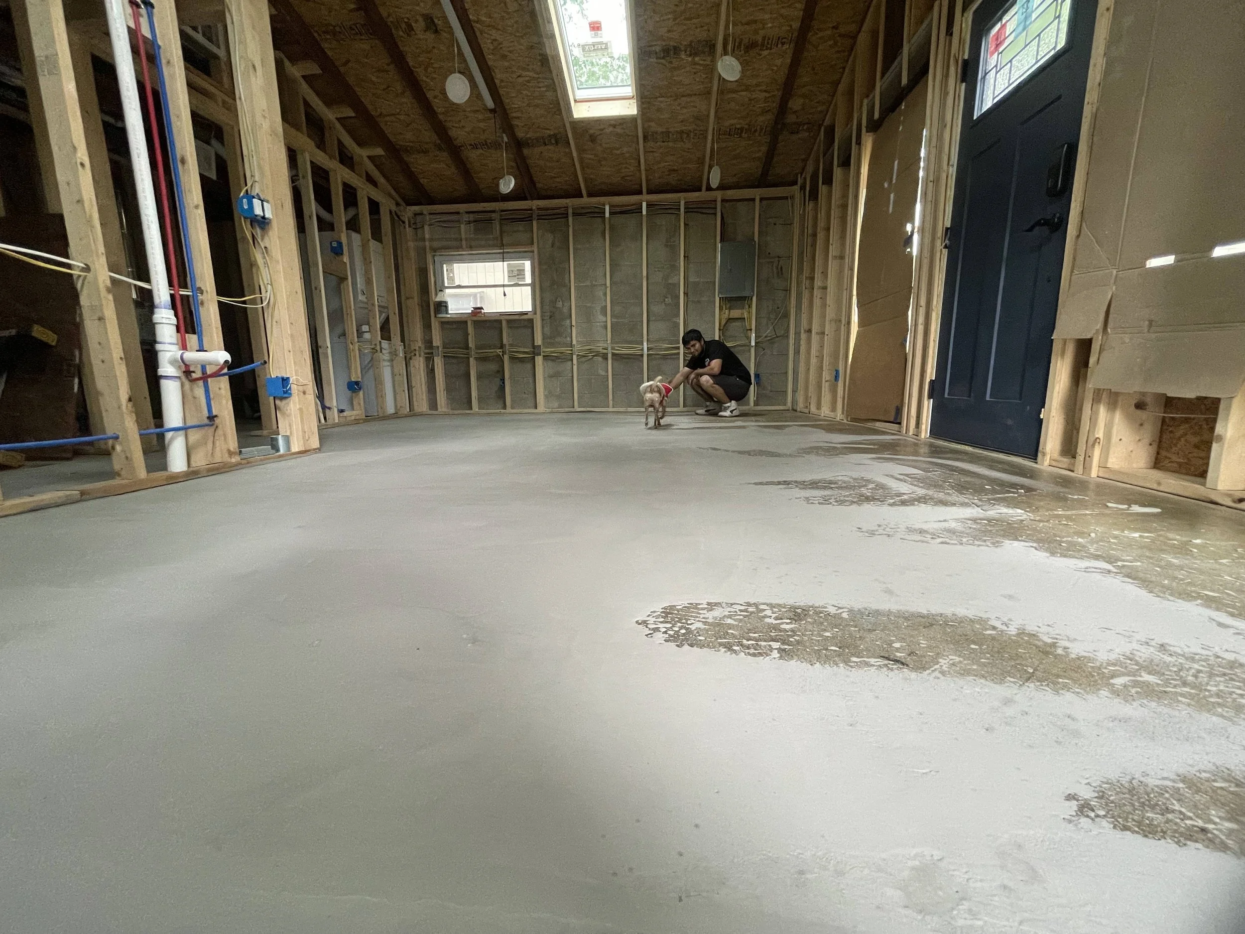A man crouching down and petting a small dog with a harness inside a partially constructed room with exposed framing, electrical wiring, and a concrete floor.
