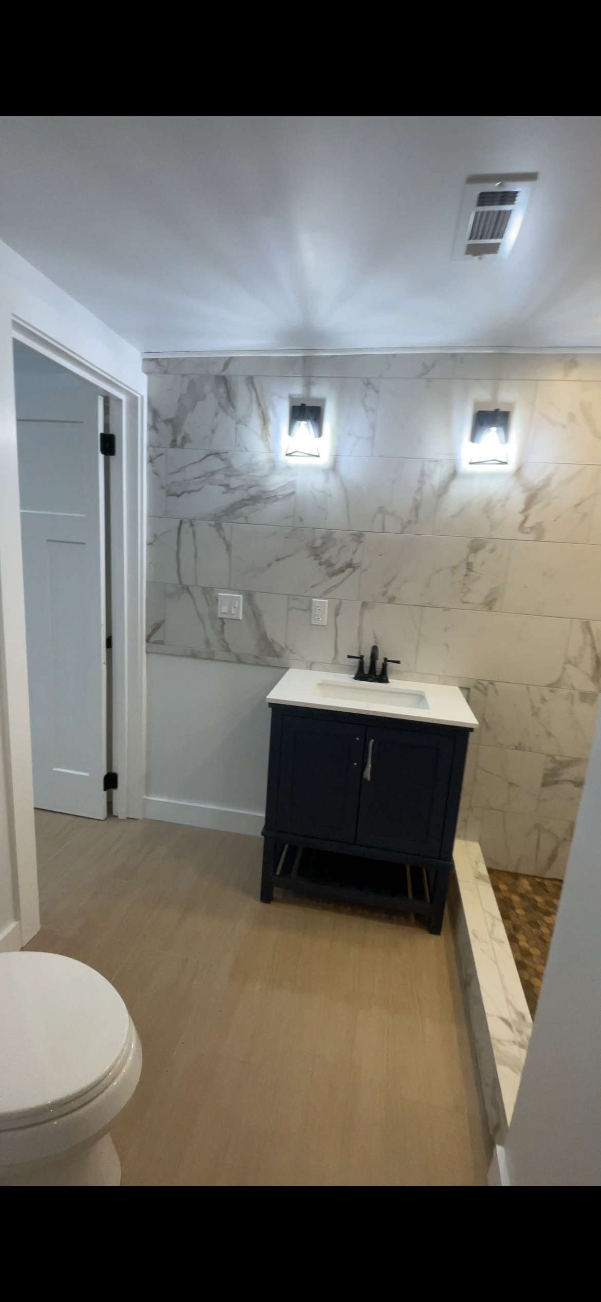 Bathroom with a white toilet, a dark blue vanity with a white sink, beige wood flooring, granite wall tiles, wall-mounted lights, and a partially visible shower area with a pebble floor.