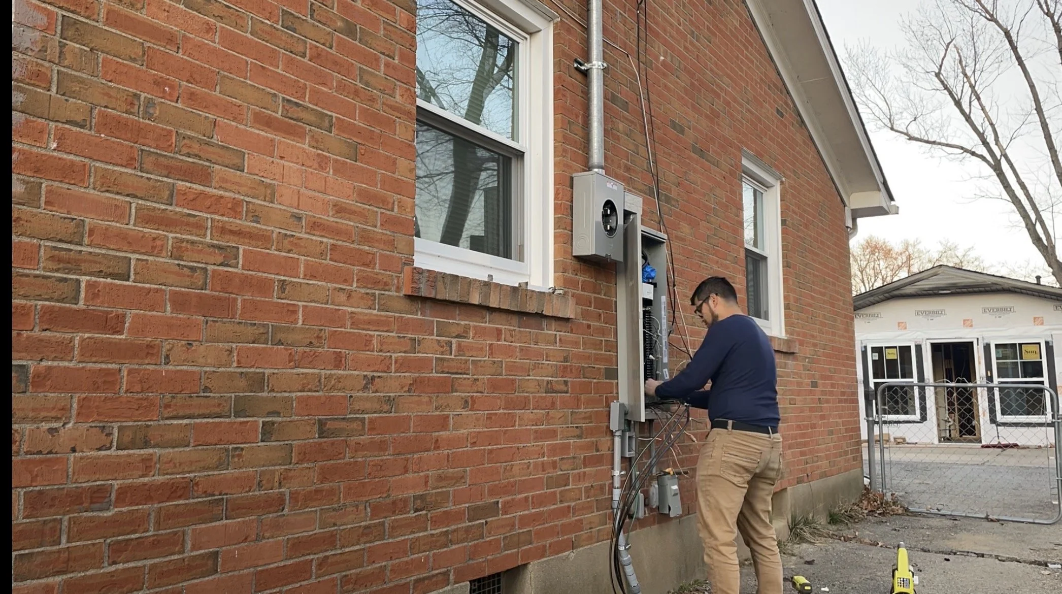 A technician working on outdoor electrical or internet cabinets on a brick house wall, with a backdrop of a new construction house and trees.