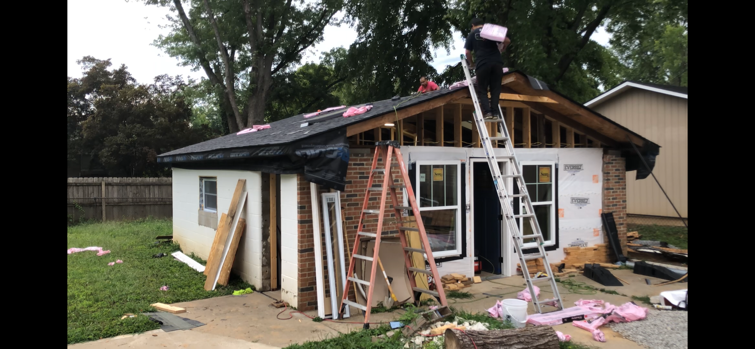 Construction workers are building or remodeling a house, with roofing in progress and various tools and materials scattered around the yard.