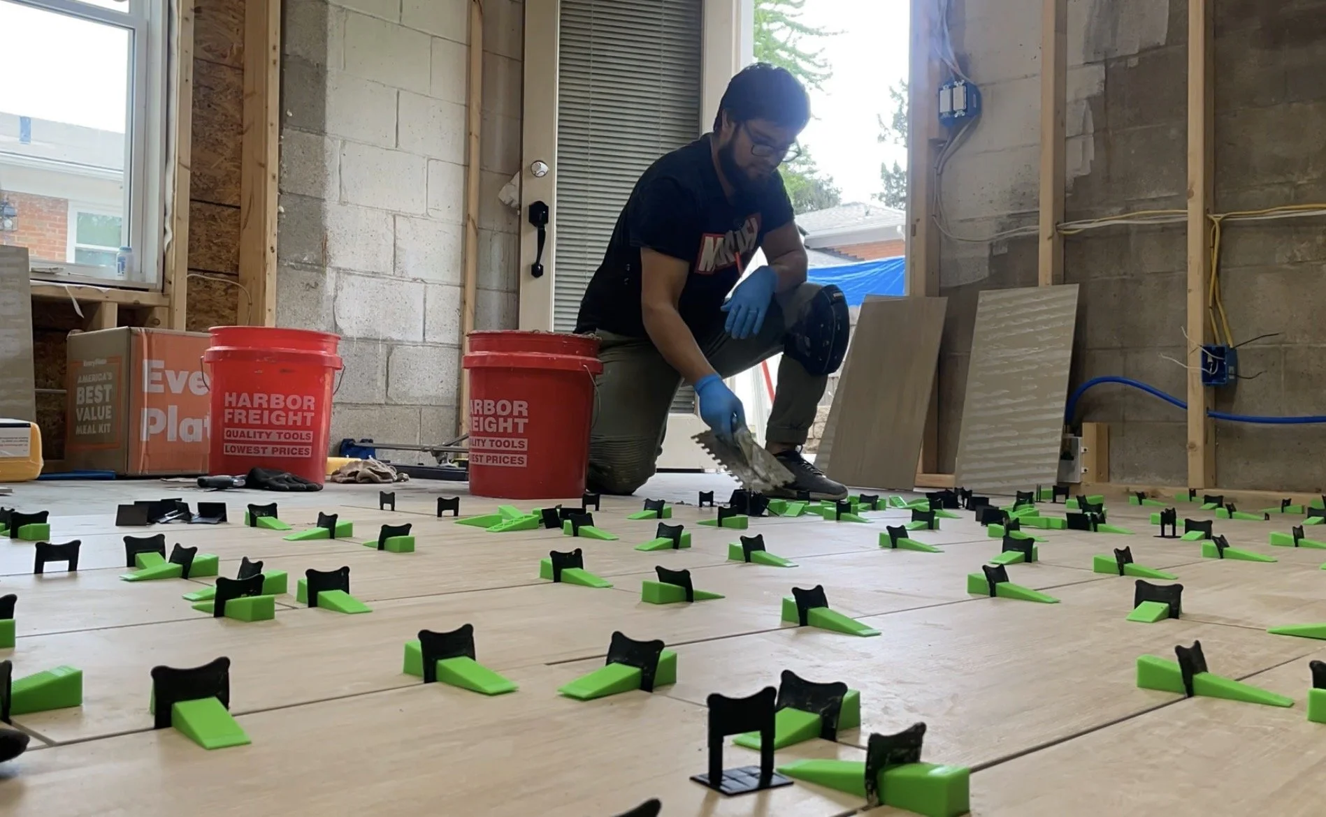 A person installing underfloor heating mats in a room with unfinished walls and large windows. The person is kneeling on the floor, wearing gloves, and using a tool to lay down the heating mats, which are black and green. There are construction tools, buckets, and boxes around.