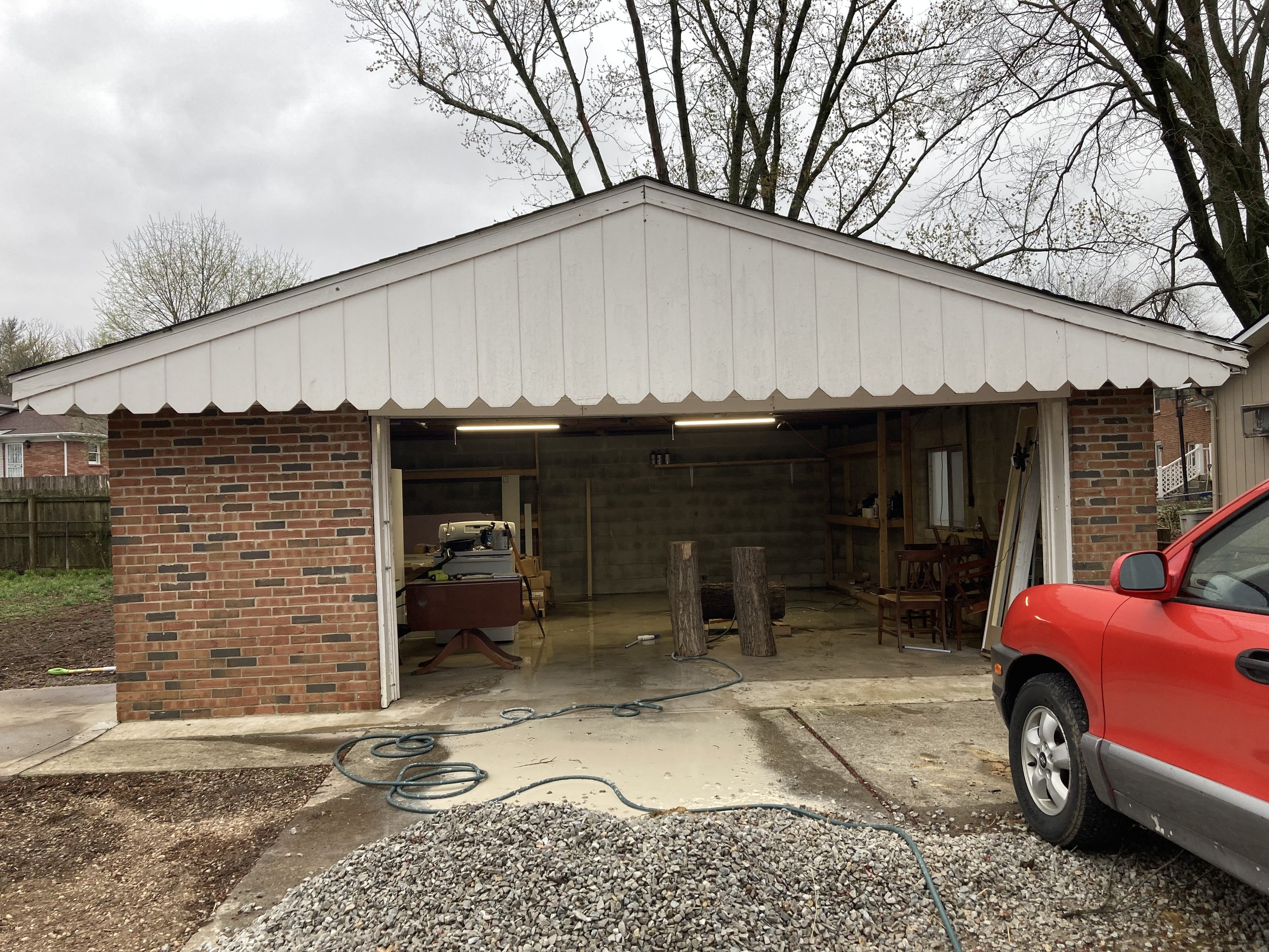 A brick garage with its door open, cluttered with tools and wooden logs inside. The roof has a white scalloped trim. A red vehicle is parked on the right, and a garden hose is on the concrete driveway. Overcast sky with leafless trees in the background.