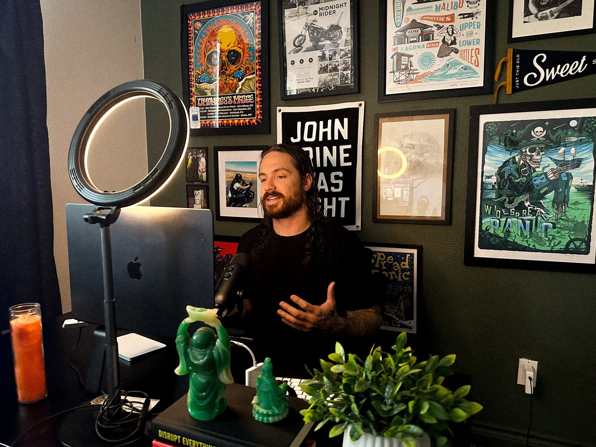 A man with long dark hair and a beard, sitting at a desk with a computer and microphone, gesturing with his hand. The background features a wall decorated with various framed posters and artwork, including a skull with a colorful hat, a motorcycle, and beach scenes. There is a ring light in the foreground, a small potted plant, and some figurines on the desk.