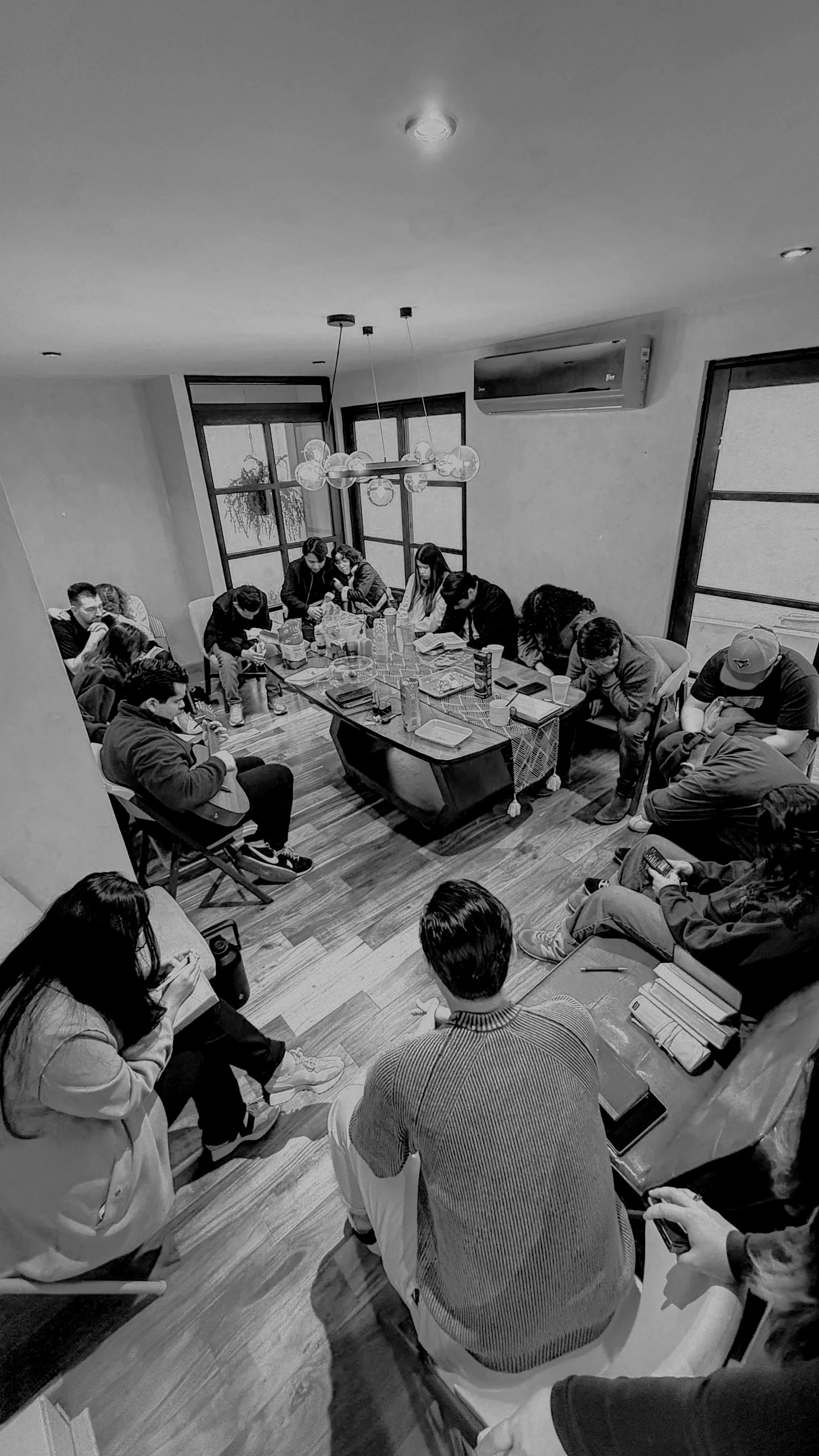 Grupo de personas reunidas en una sala, algunas leyendo libros o usando teléfonos, una en la esquina tocando guitarra, en una mesa hay comida y bebidas, ventanas grandes y aire acondicionado.