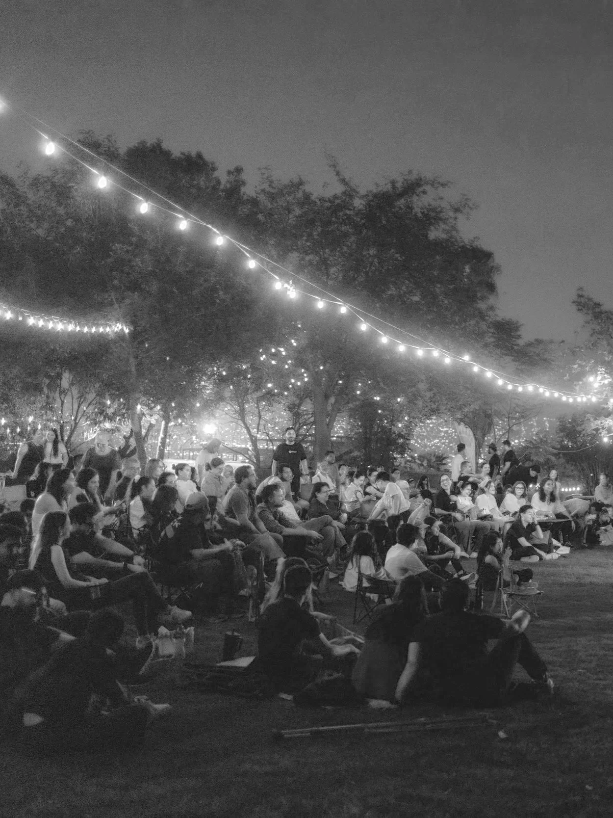 Reunión al aire libre durante la noche con muchas personas sentadas en sillas y en el suelo, decoraciones con luces en árboles y cables iluminados, ambiente festivo o de evento comunitario.
