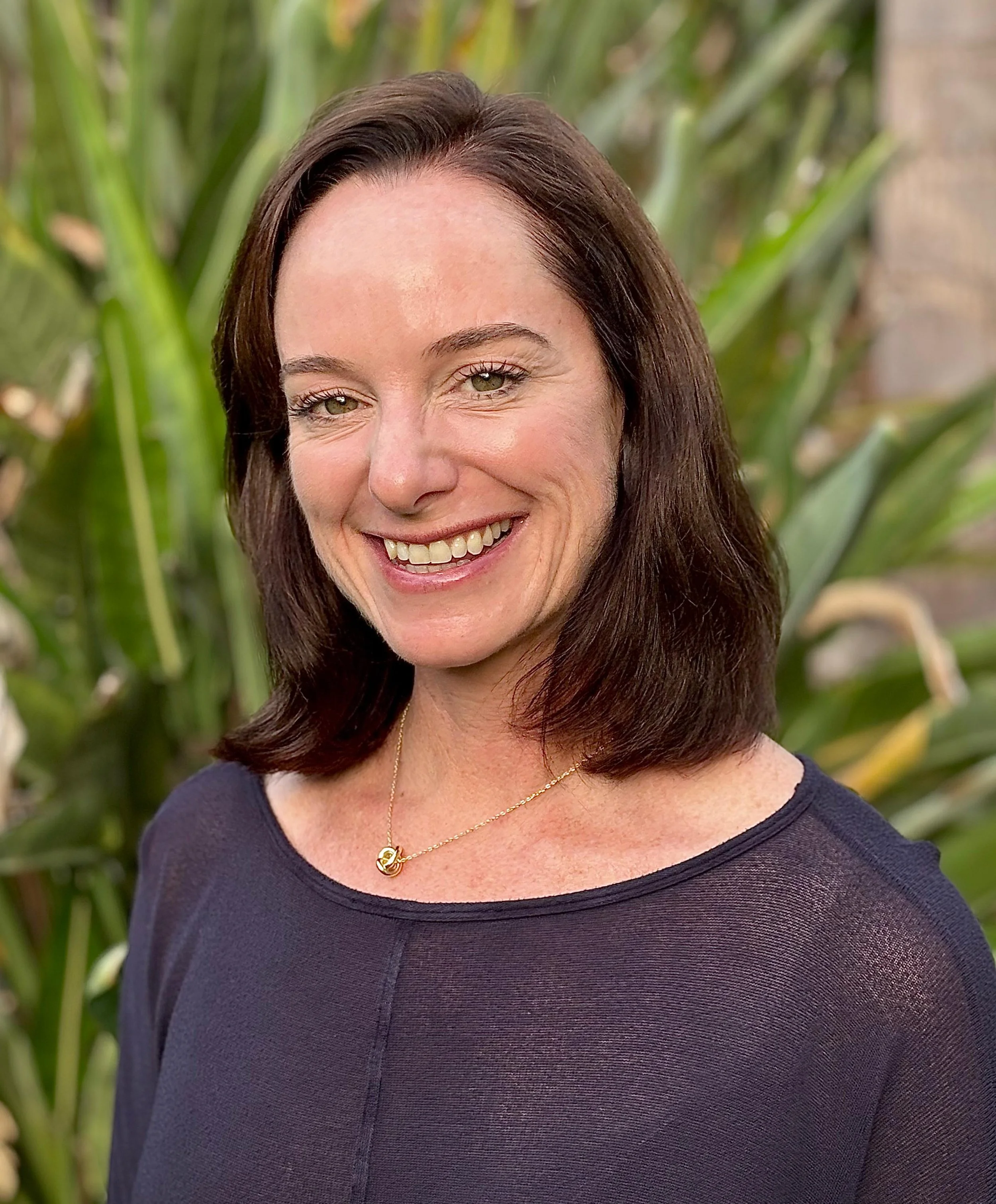 Close-up of a woman smiling outdoors with green plants in the background, wearing a dark top and a gold necklace.
