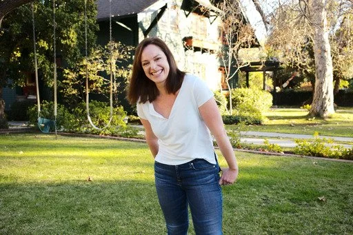 A young woman smiling and standing outdoors on a green lawn with trees and houses in the background.