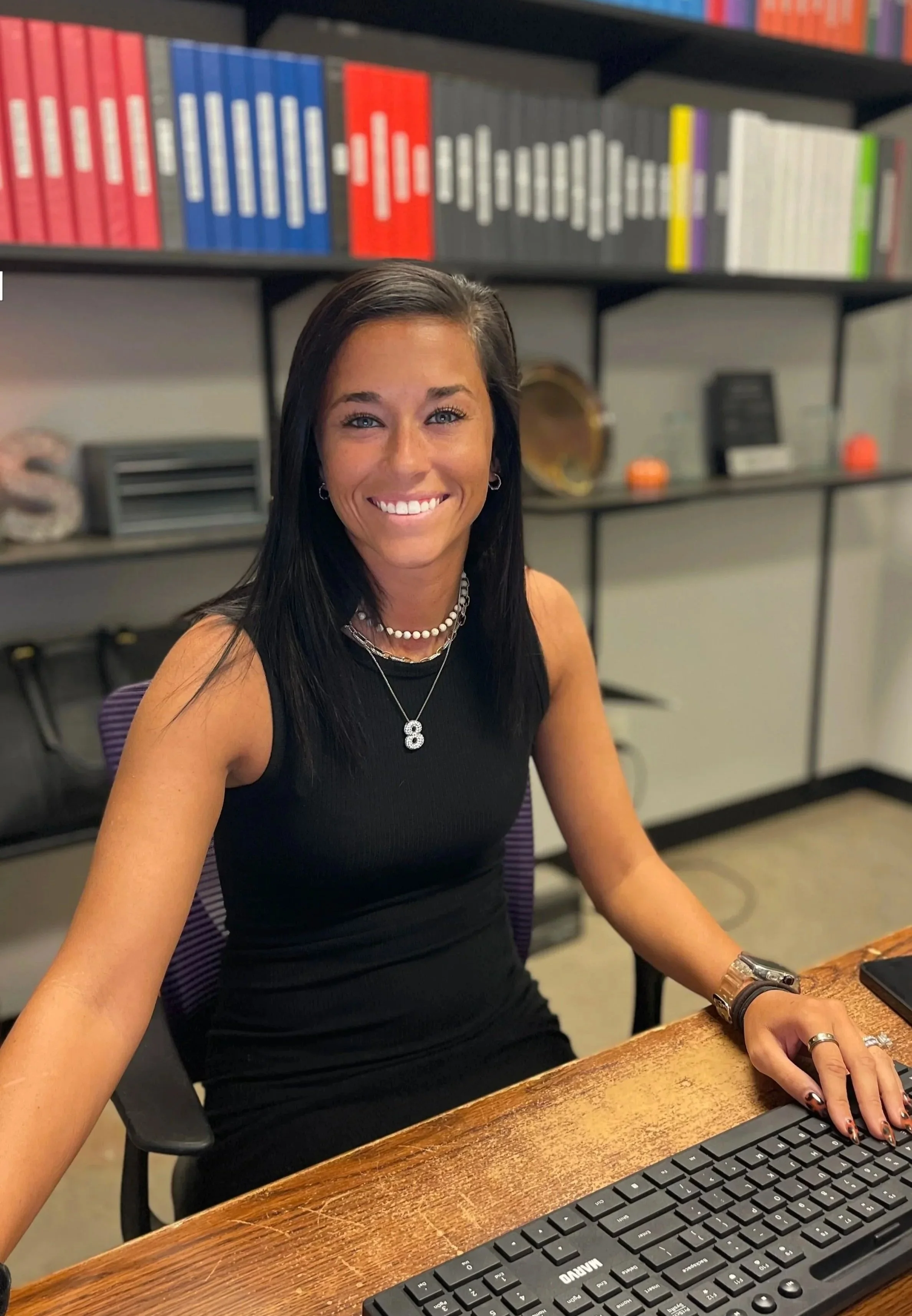 A woman with long dark hair smiling at the camera, sitting at a wooden desk with a computer keyboard in front of her. She is wearing a black sleeveless top, a pearl necklace, and jewelry on her wrist and fingers. Behind her is a bookshelf with colorful binders and other items.