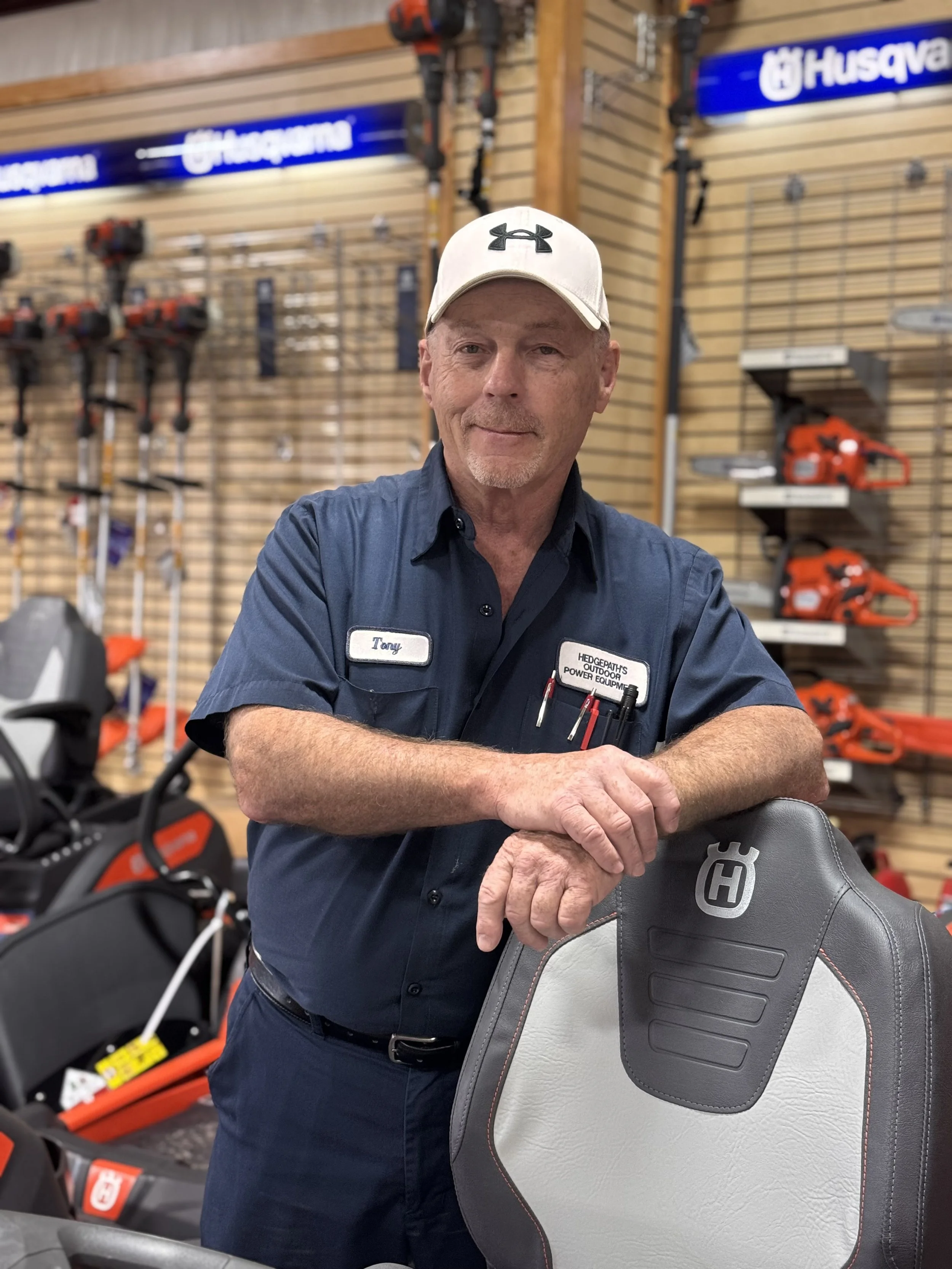 A man with a name badge that says Tony, wearing a white cap with the Under Armour logo, standing inside a store that sells outdoor power equipment and chainsaws, with chainsaws displayed on a wooden wall in the background.