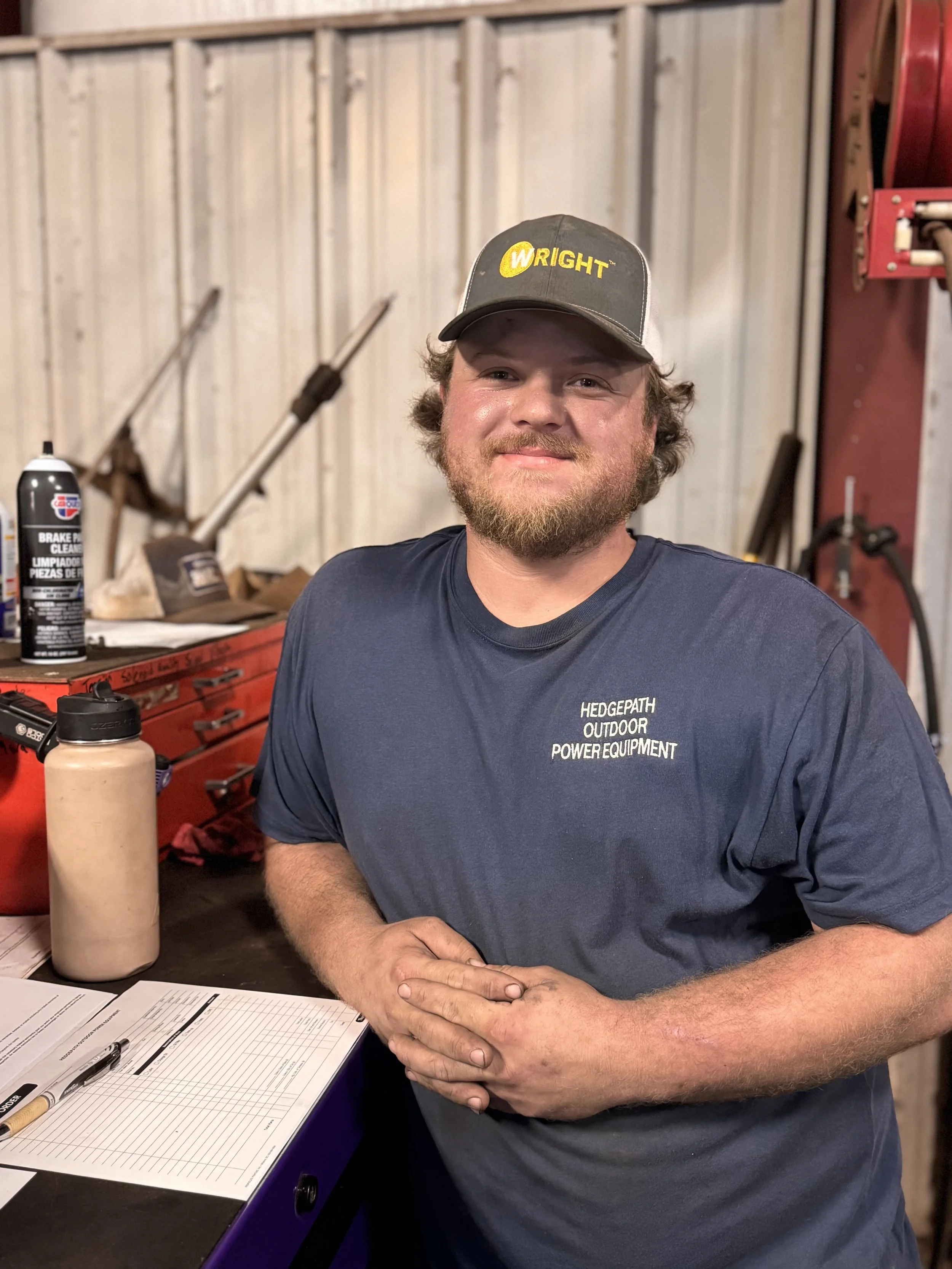A man with a beard wearing a gray baseball cap with yellow lettering, sitting at a workspace with papers and a pen, in a workshop or garage with tools and a red tool chest in the background.
