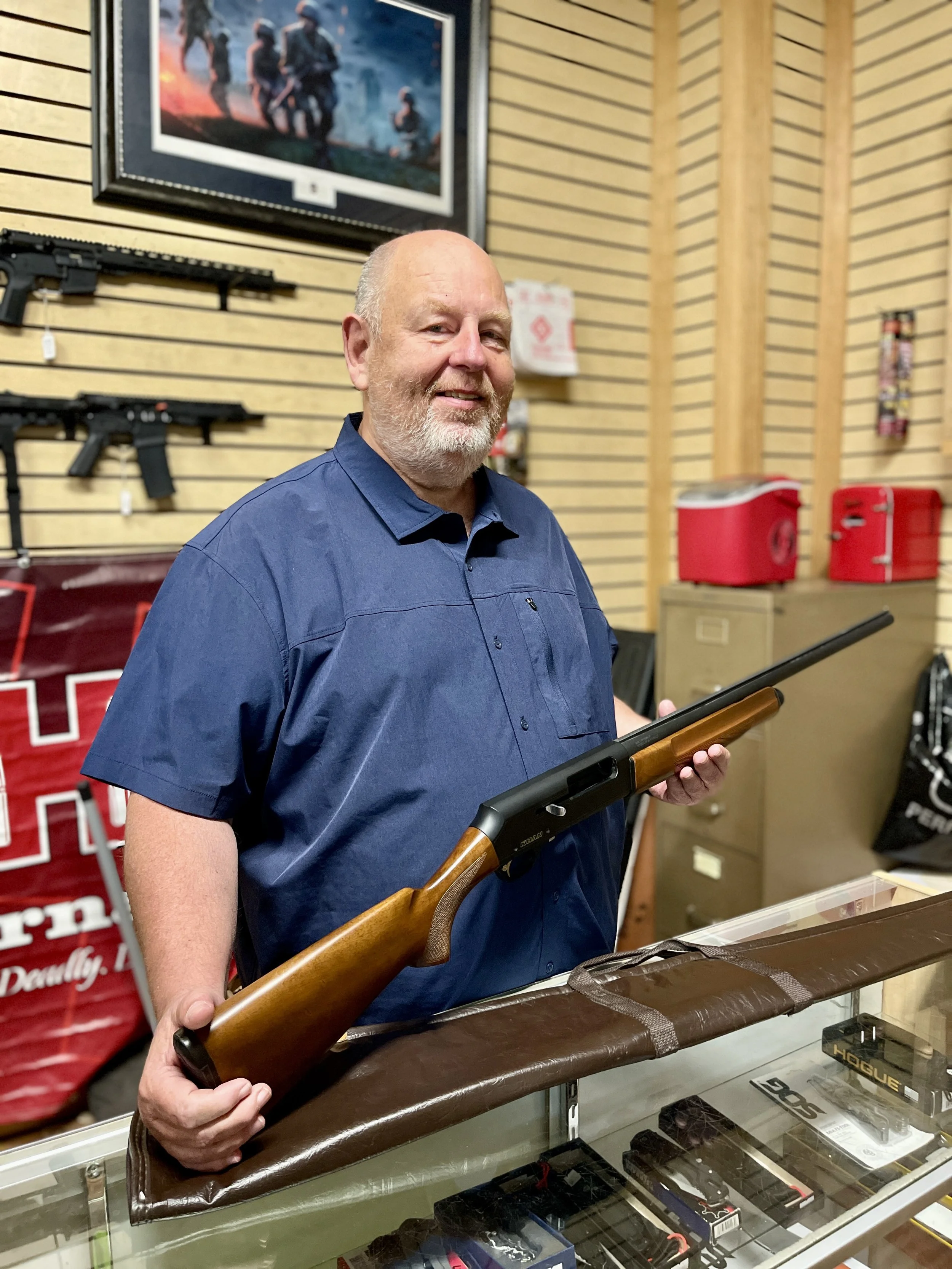 An older man with a beard and blue shirt holding a rifle inside a store with firearms on the wall behind him.