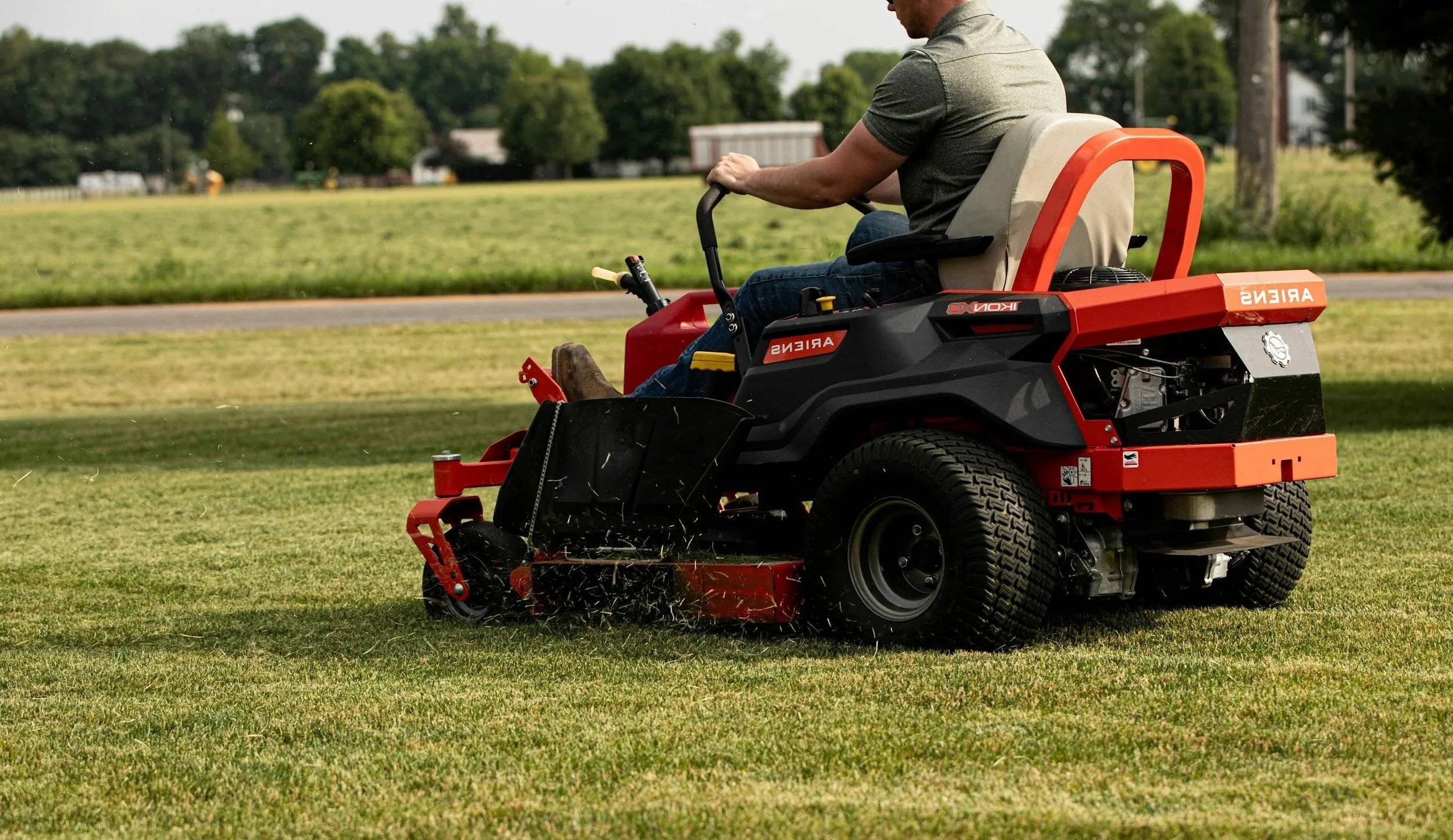 A person in a gray shirt and jeans operating a Kubota Ariens riding lawn mower on a grassy field, with trees and houses in the background.