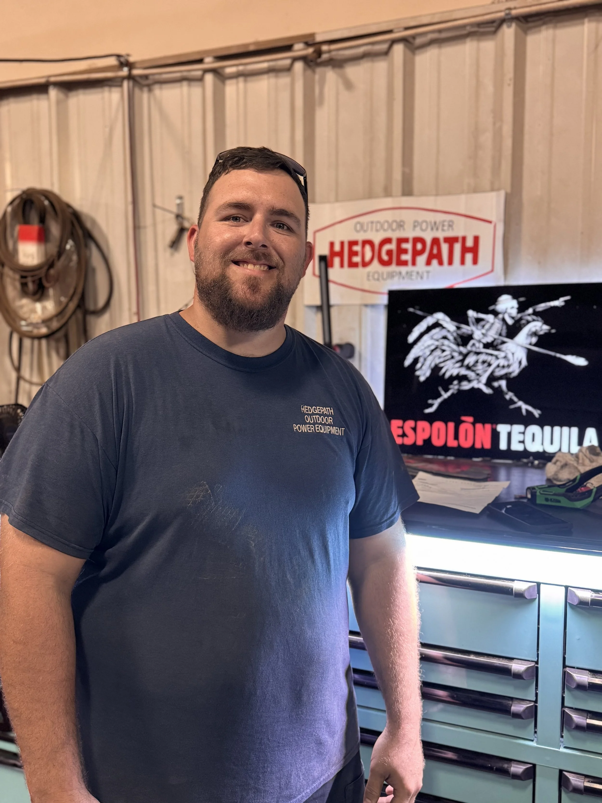 A smiling man with a beard and short dark hair, wearing a dark t-shirt, standing in front of a workbench with drawers, a computer monitor, and tool hooks, indoors with wooden wall paneling and a sign for Hedgepath Outdoor Power Equipment.