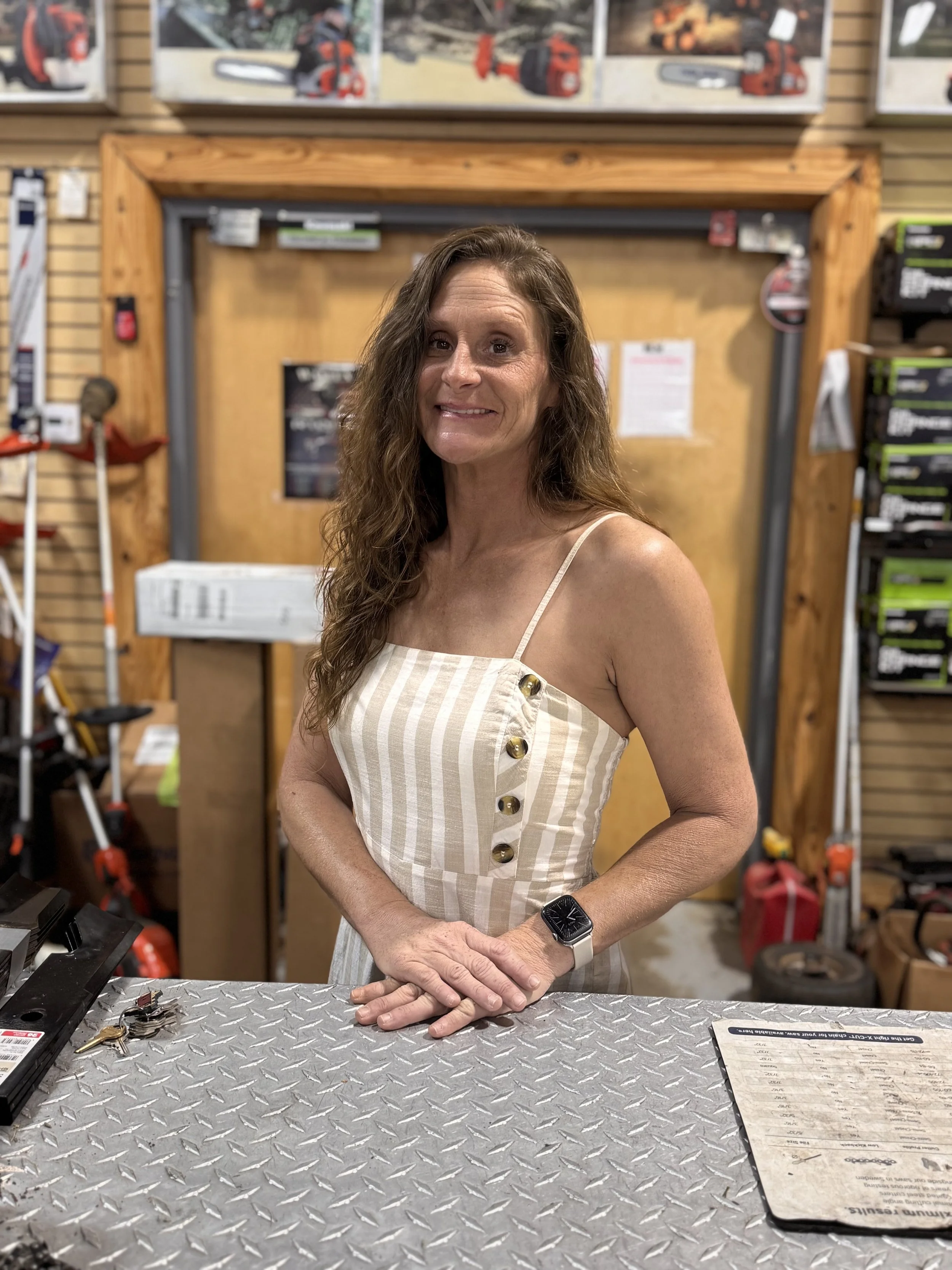 A woman with long brown hair smiling, wearing a light striped dress with buttons on the side, standing behind a metal counter in a hardware store with tools and equipment around.