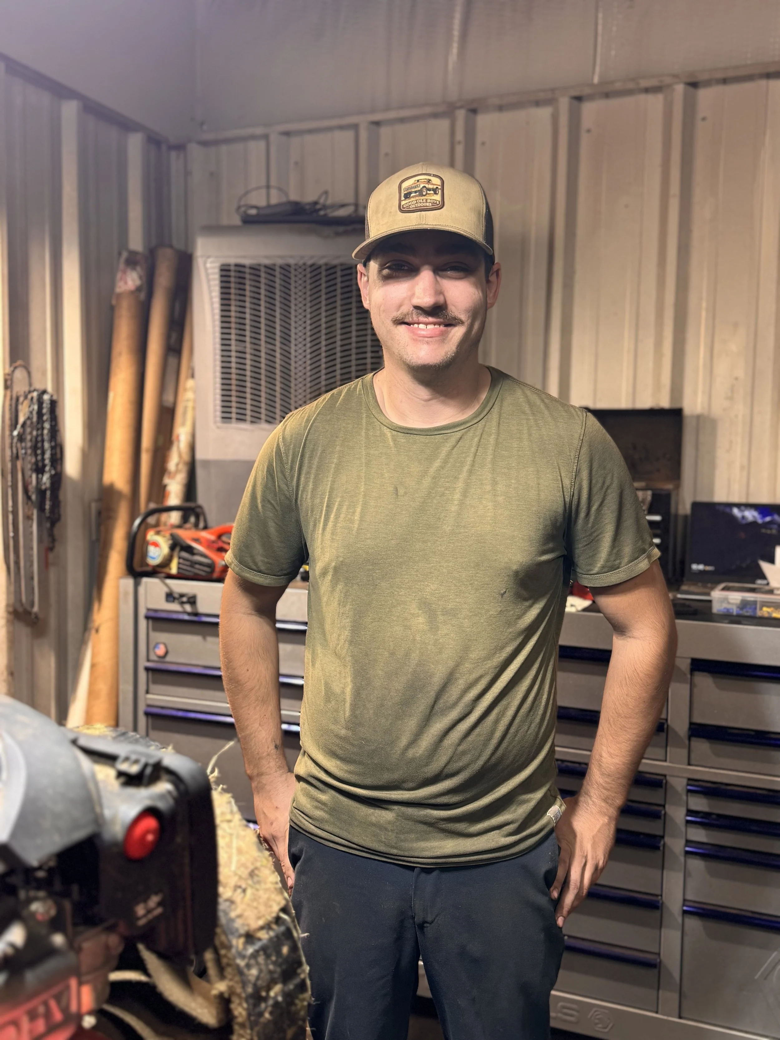 A man in a green t-shirt and dark pants poses smiling in a workshop with tools and equipment behind him, including long-handled tools, a small drill, and a large storage cabinet.