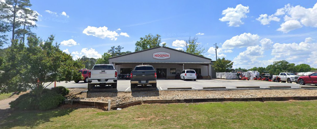Parking lot in front of a metal building with a sign that reads 'Hedgepath', surrounded by trees and blue sky with clouds.