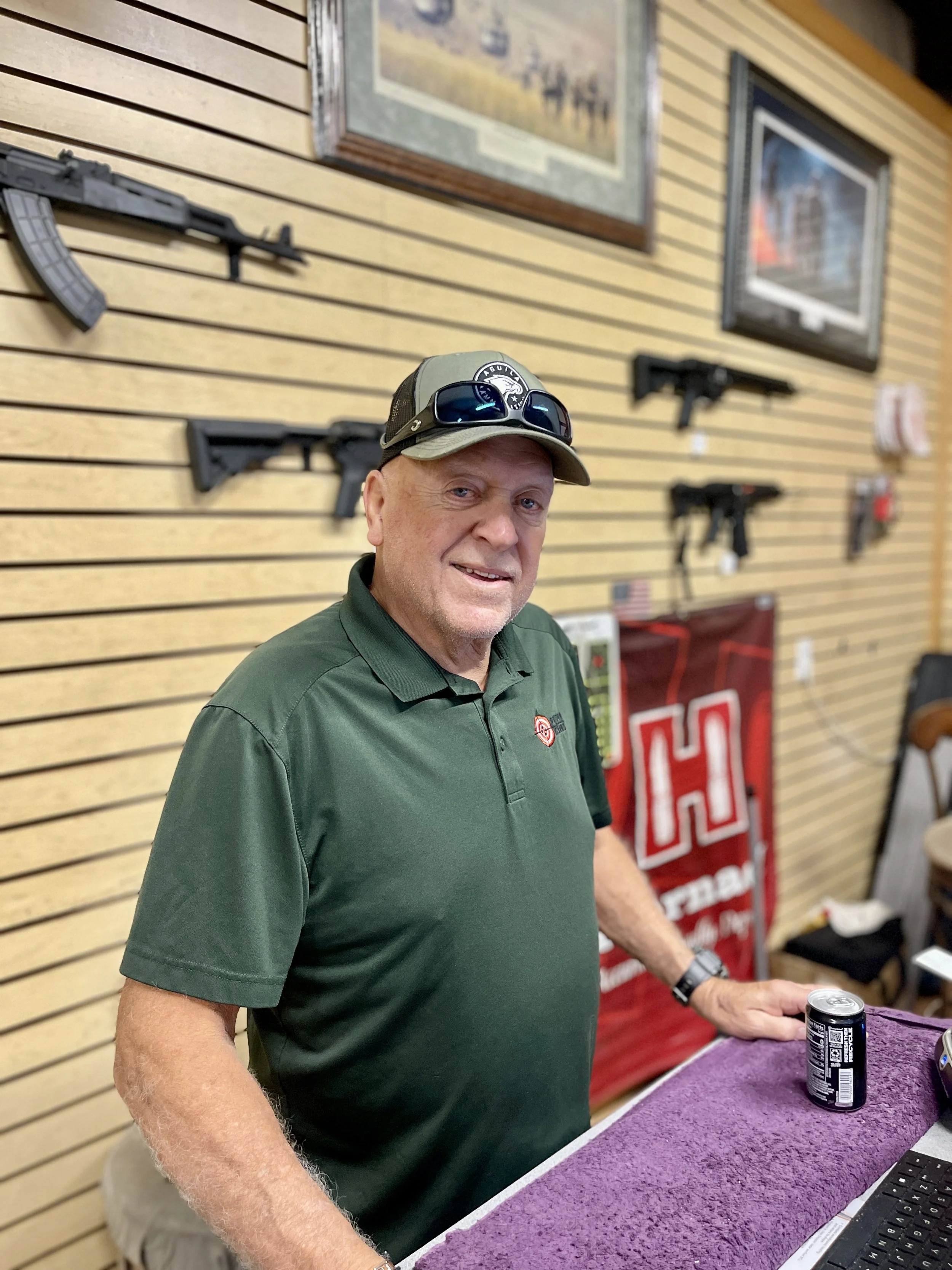 A man wearing a green polo shirt, cap, and sunglasses on his head stands behind a purple counter, with a wall of firearms and framed pictures behind him.