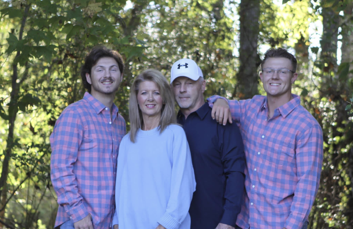 Four people standing outdoors in a wooded area, smiling at the camera. Two men with plaid shirts, a woman in a light blue top, and a man in a dark shirt and white cap.