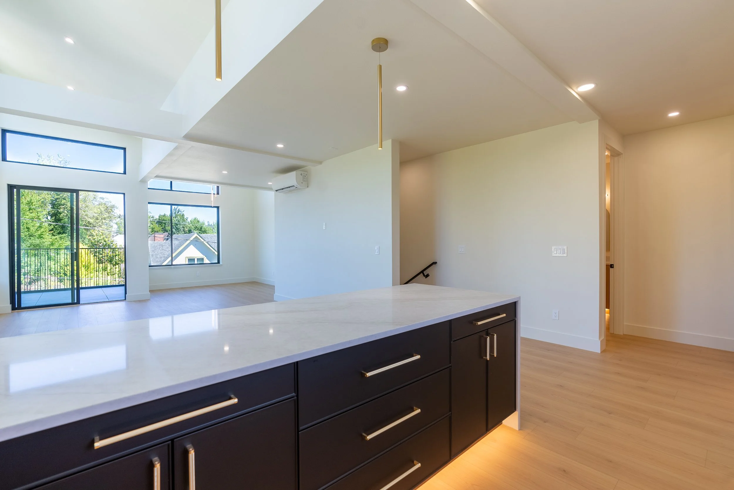 Modern kitchen with dark cabinets, white countertop, large windows, hardwood floors, and a view of a house and trees outside.