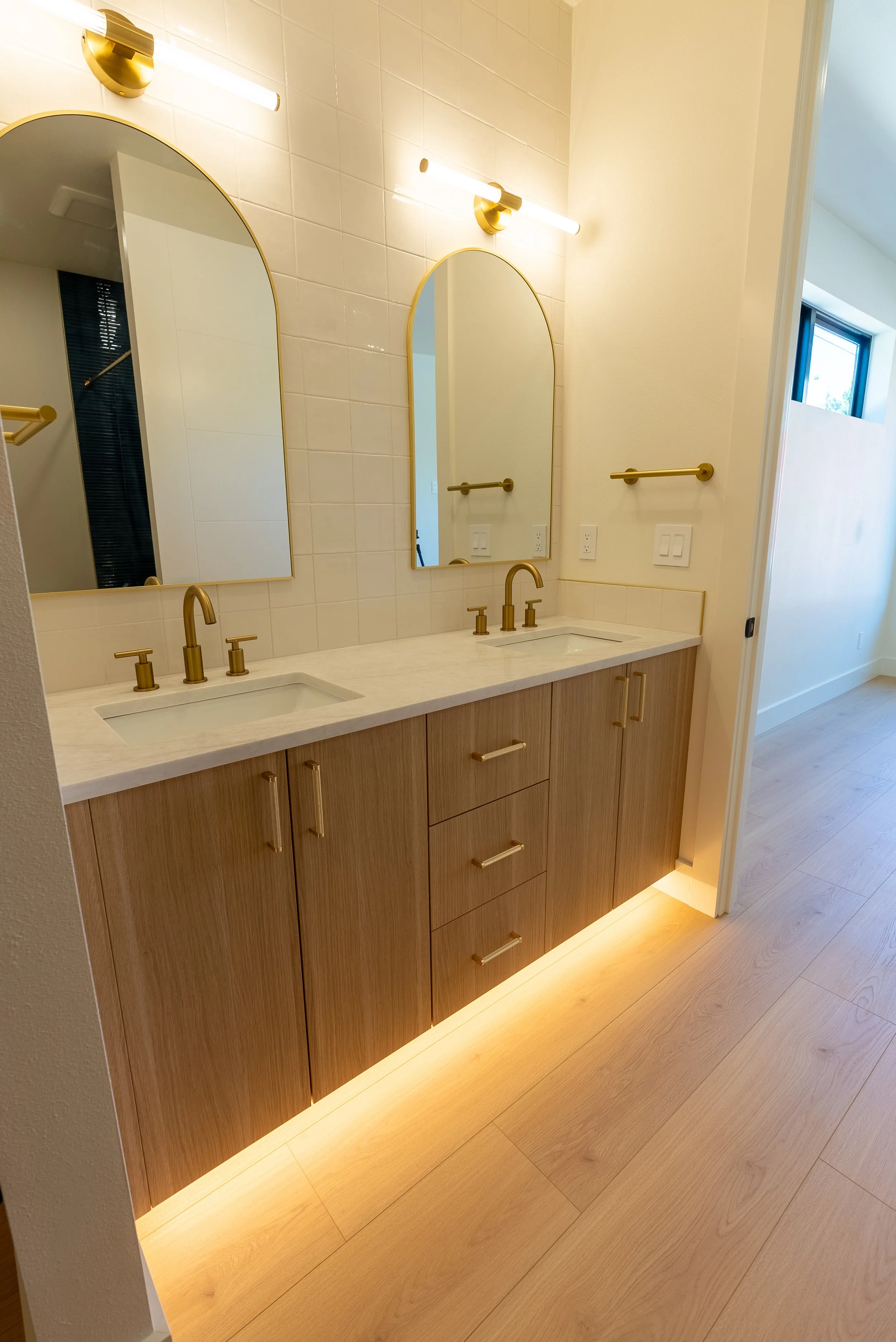 Modern bathroom vanity with two pops of gold fixtures and a backlit wooden base, pairing a white countertop with double sinks, gold faucet, and large mirrors. Light-colored wooden flooring.