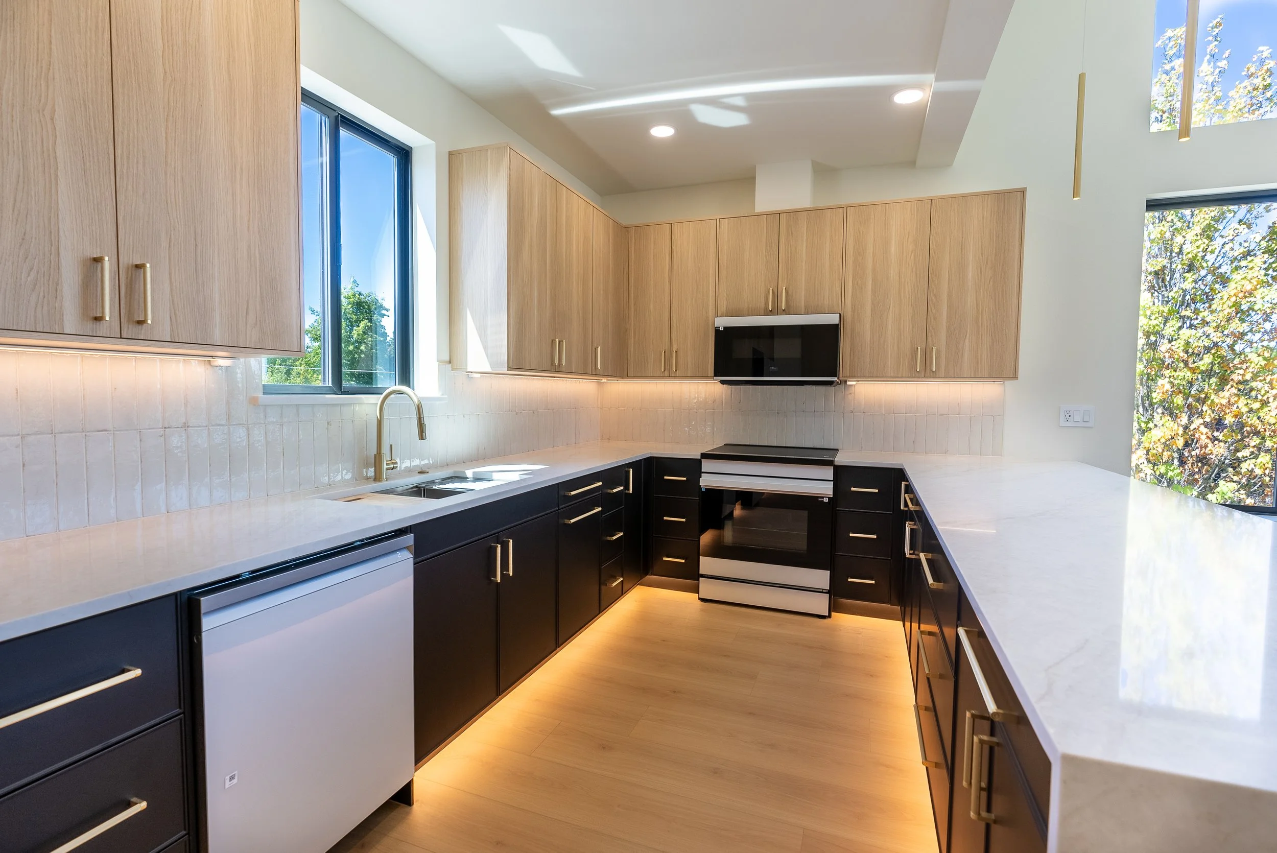 Modern kitchen with wooden upper cabinets, black lower cabinets, white countertops, a window with a view of trees, and built-in appliances including a microwave and oven.