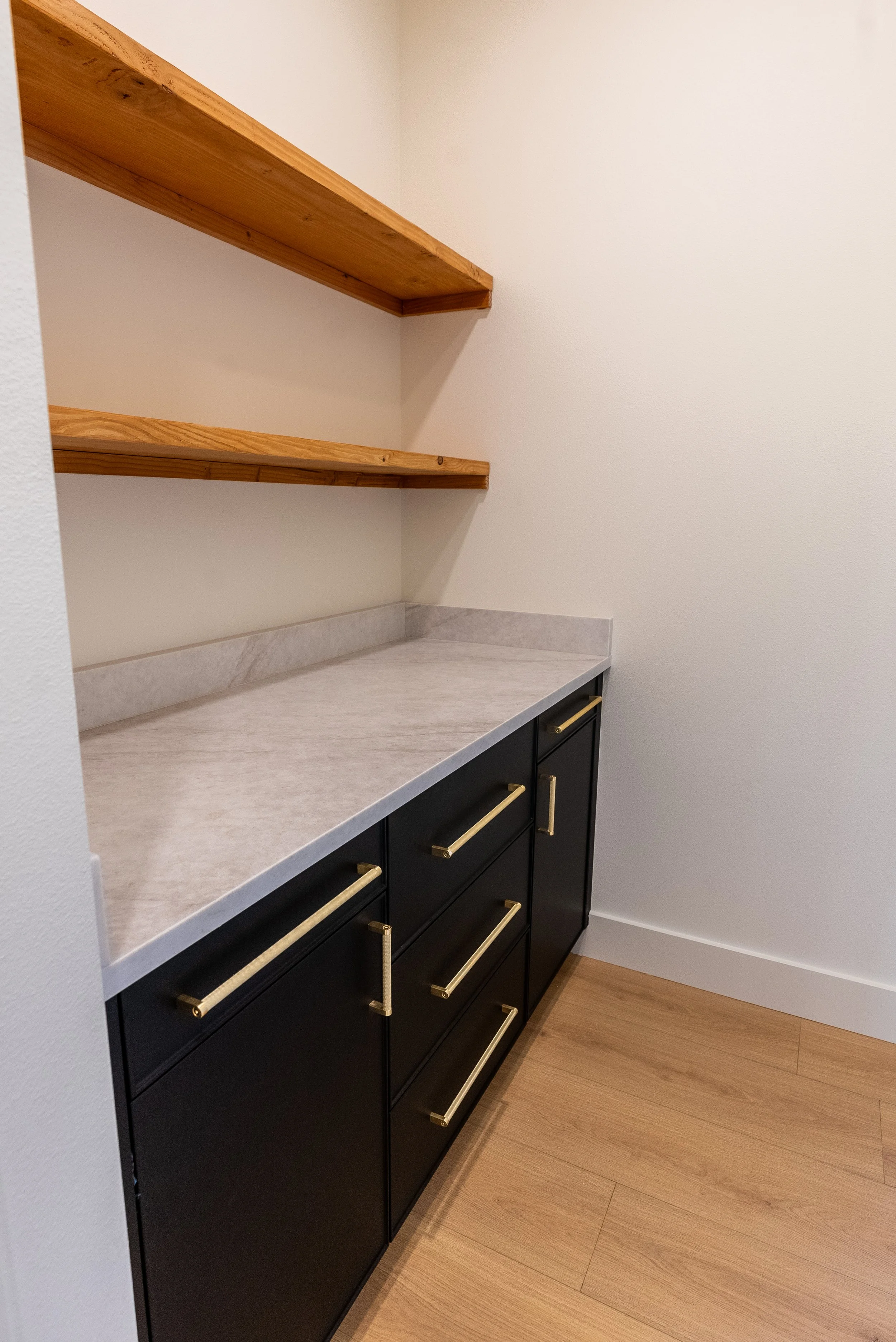 A kitchen corner featuring a white marble countertop, black cabinets with gold handles, and wooden open shelves mounted on a white wall, with wooden flooring below.