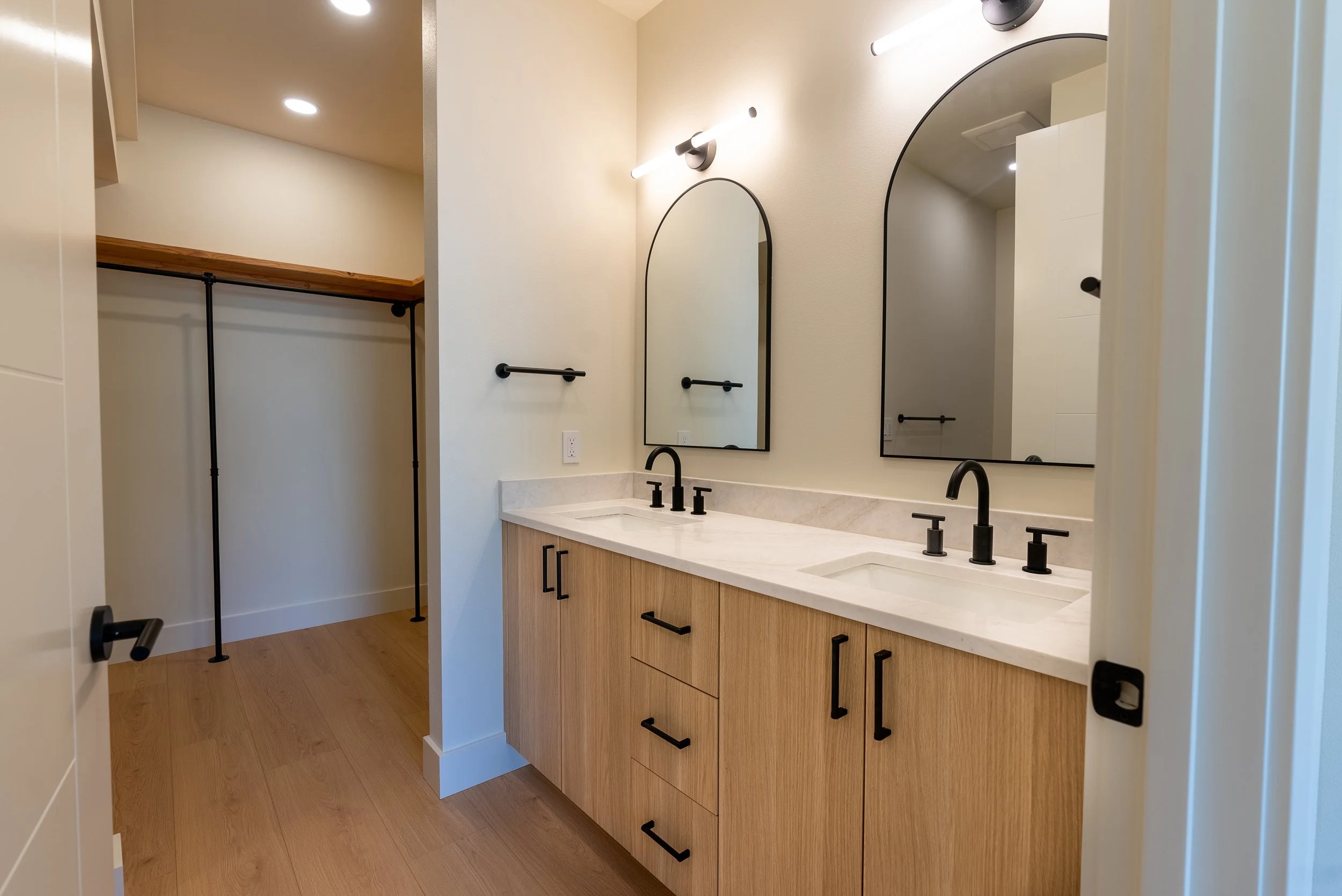 Modern bathroom vanity with a light wooden cabinet, black handles, a white countertop, and double black sinks with matching black faucets. Two tall mirrors with black frames and a white wall with two black light fixtures are visible. The room has lig