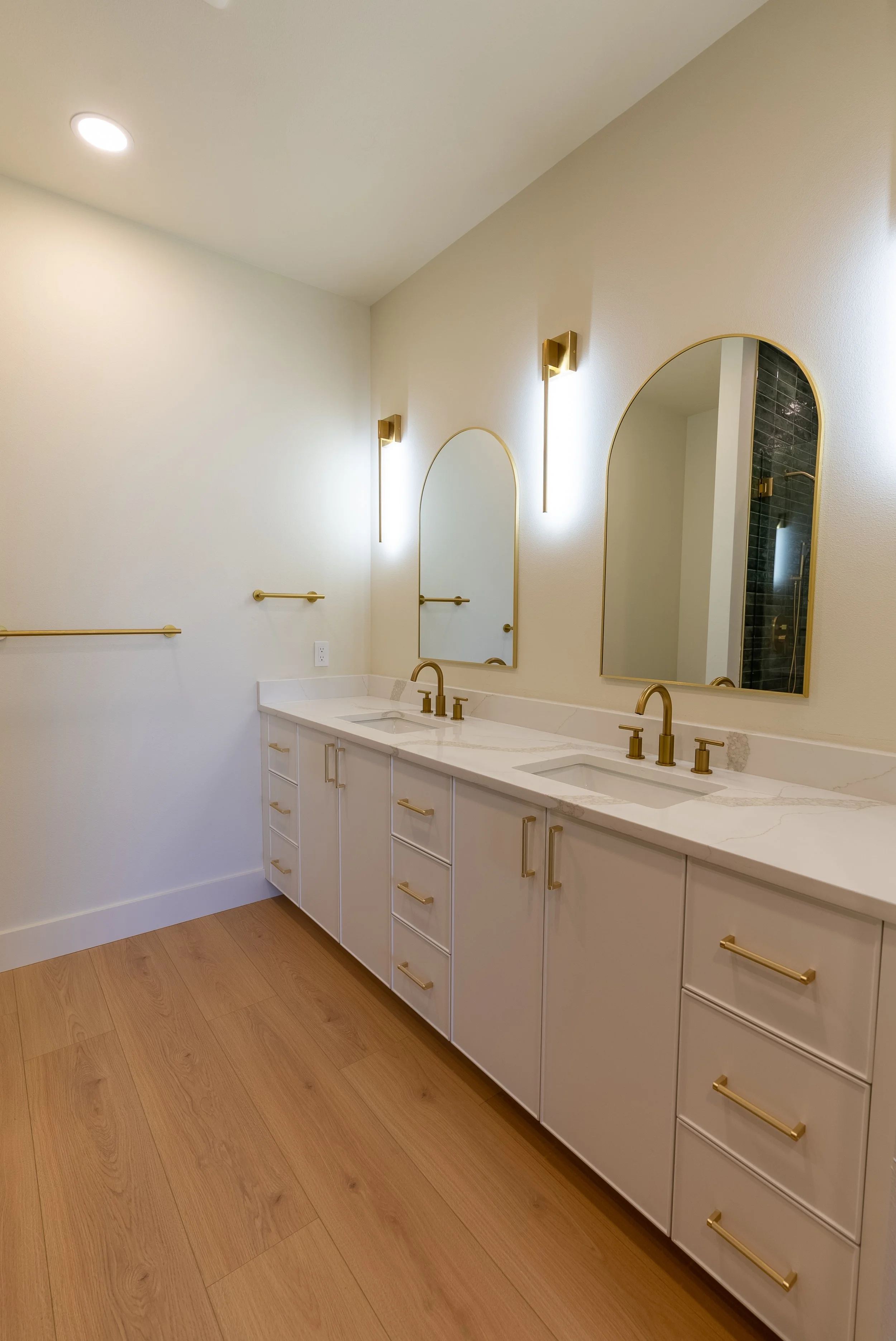 Bathroom vanity with white cabinets, gold handles, a marble countertop, two arched mirrors, and gold fixtures. The wall has gold sconces and towel bars, and there are wood floors.