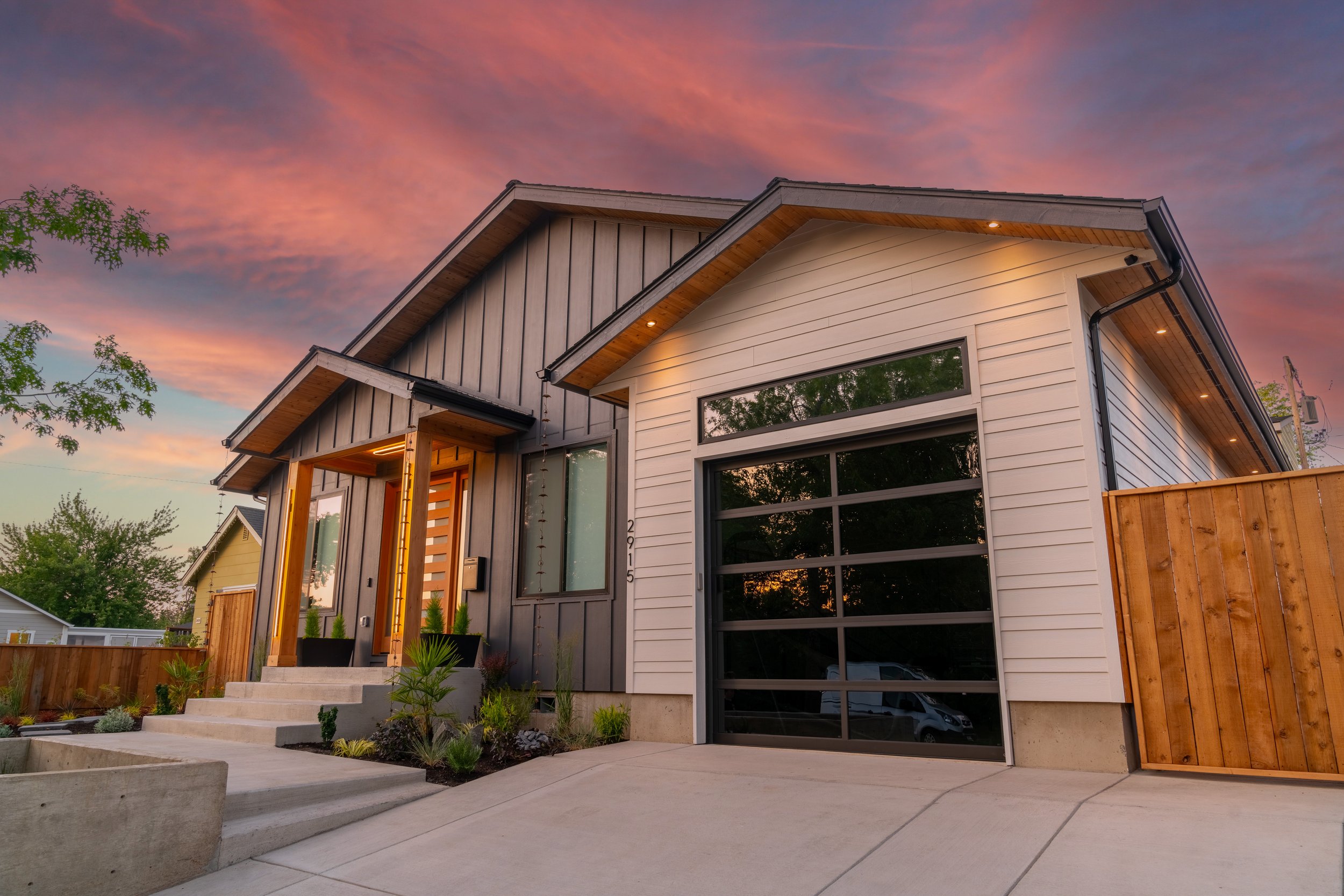 Modern house with large windows, white and gray exterior, wooden accents under the roof, front steps leading to the entrance, a garden with plants, wooden fence, and a colorful sunset sky.